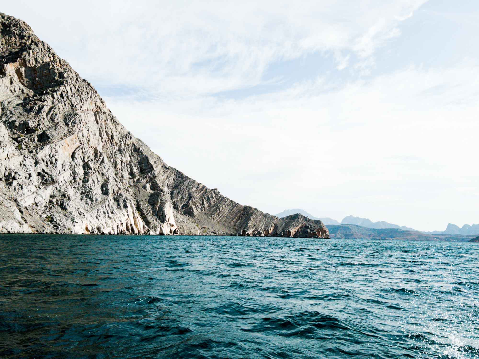 Rocky coastal landscape with clear blue water and distant mountains under a bright sky.
