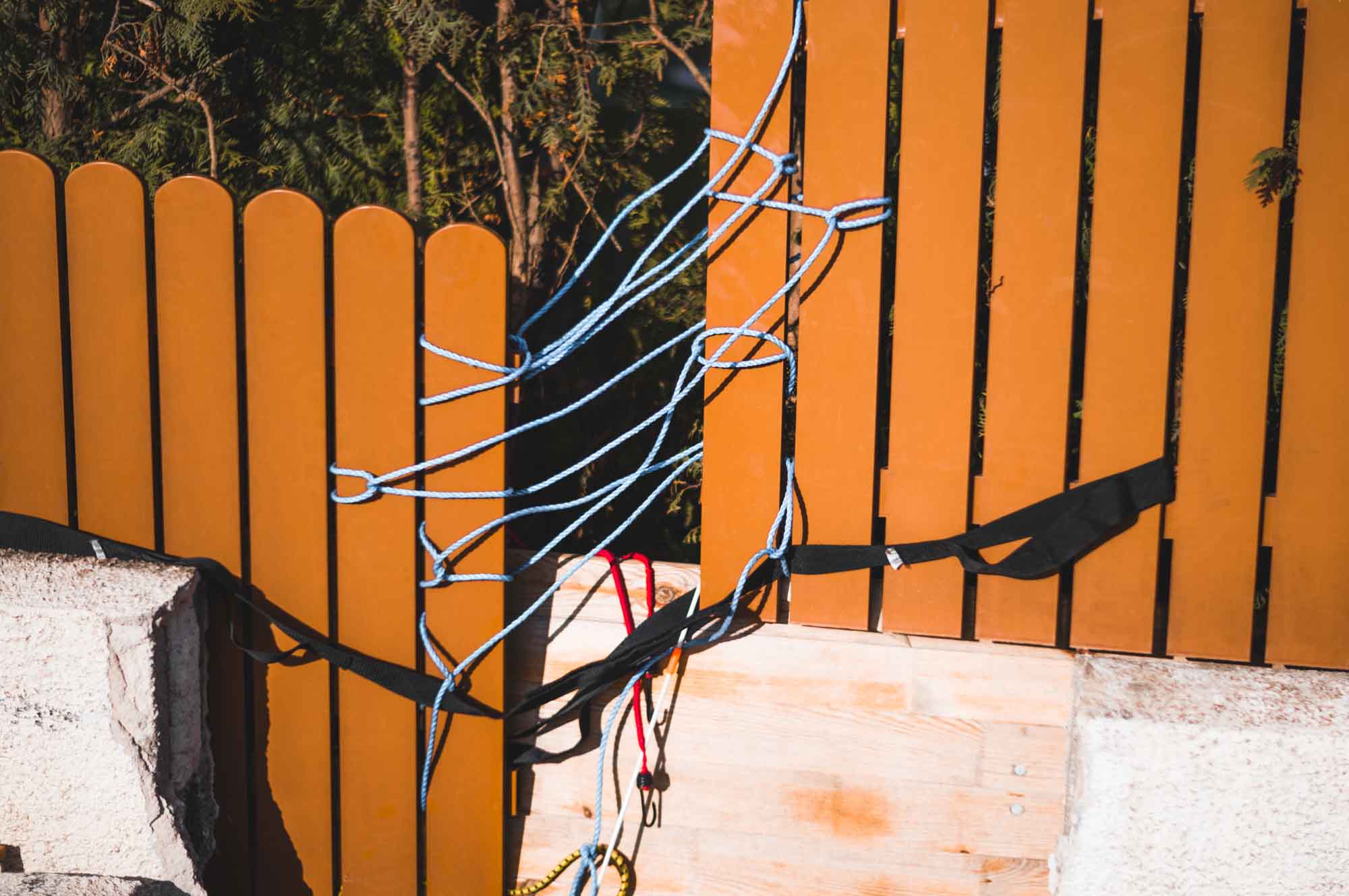 Wooden fence with blue rope and black straps tied, DIY outdoor project in sunlight.