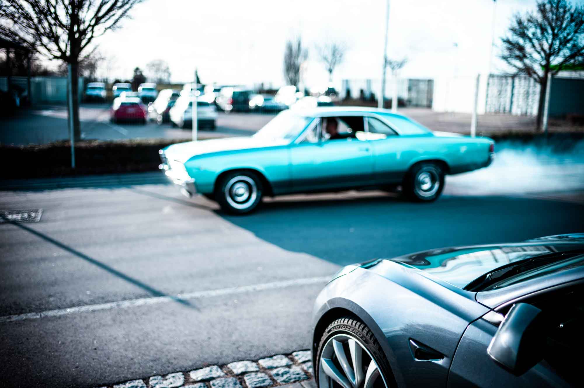 Classic blue car skids on street, leaving smoke trail on sunny day, with modern car in foreground.