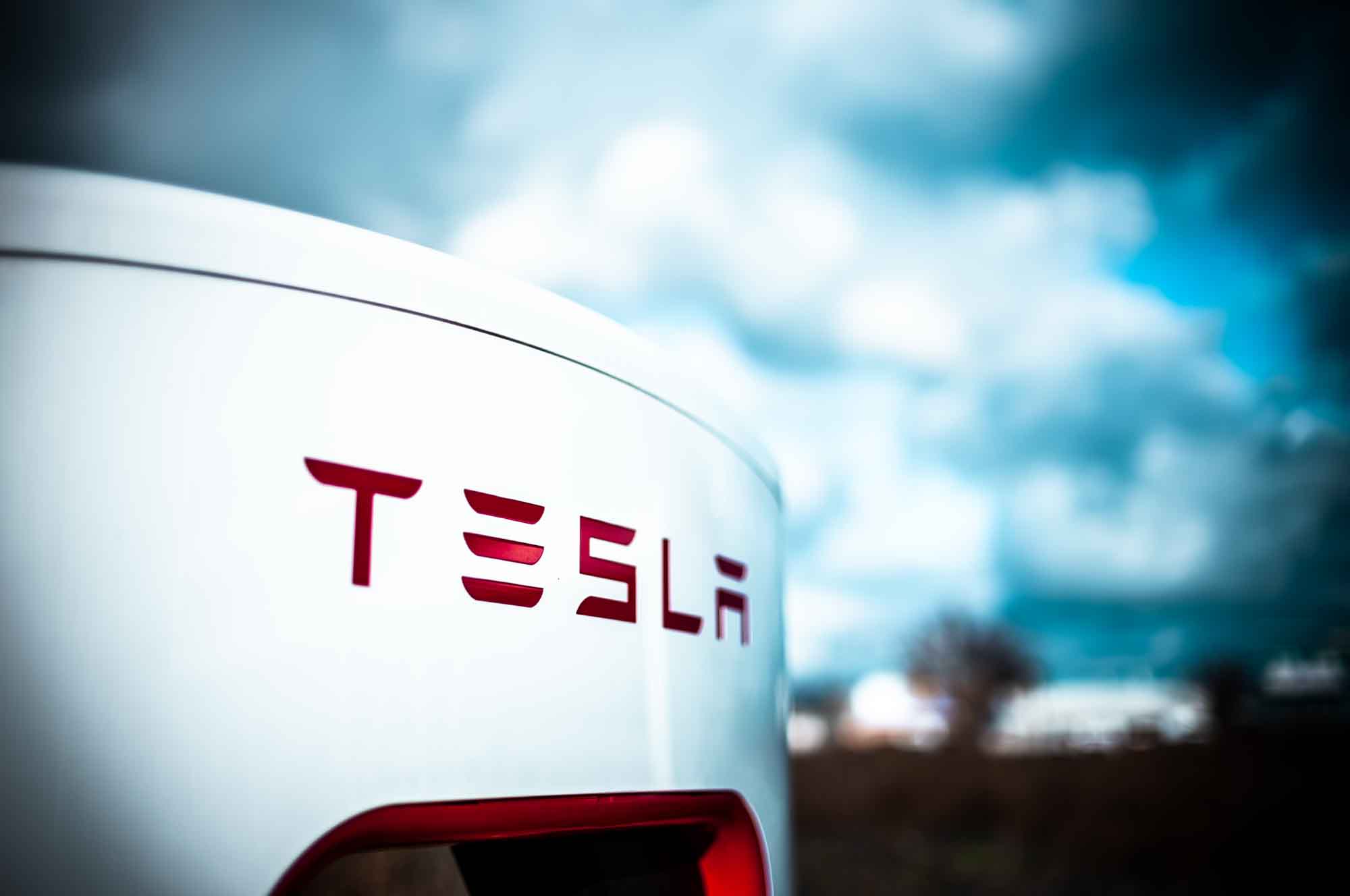 Close-up of Tesla logo on a charging station under a cloudy sky.