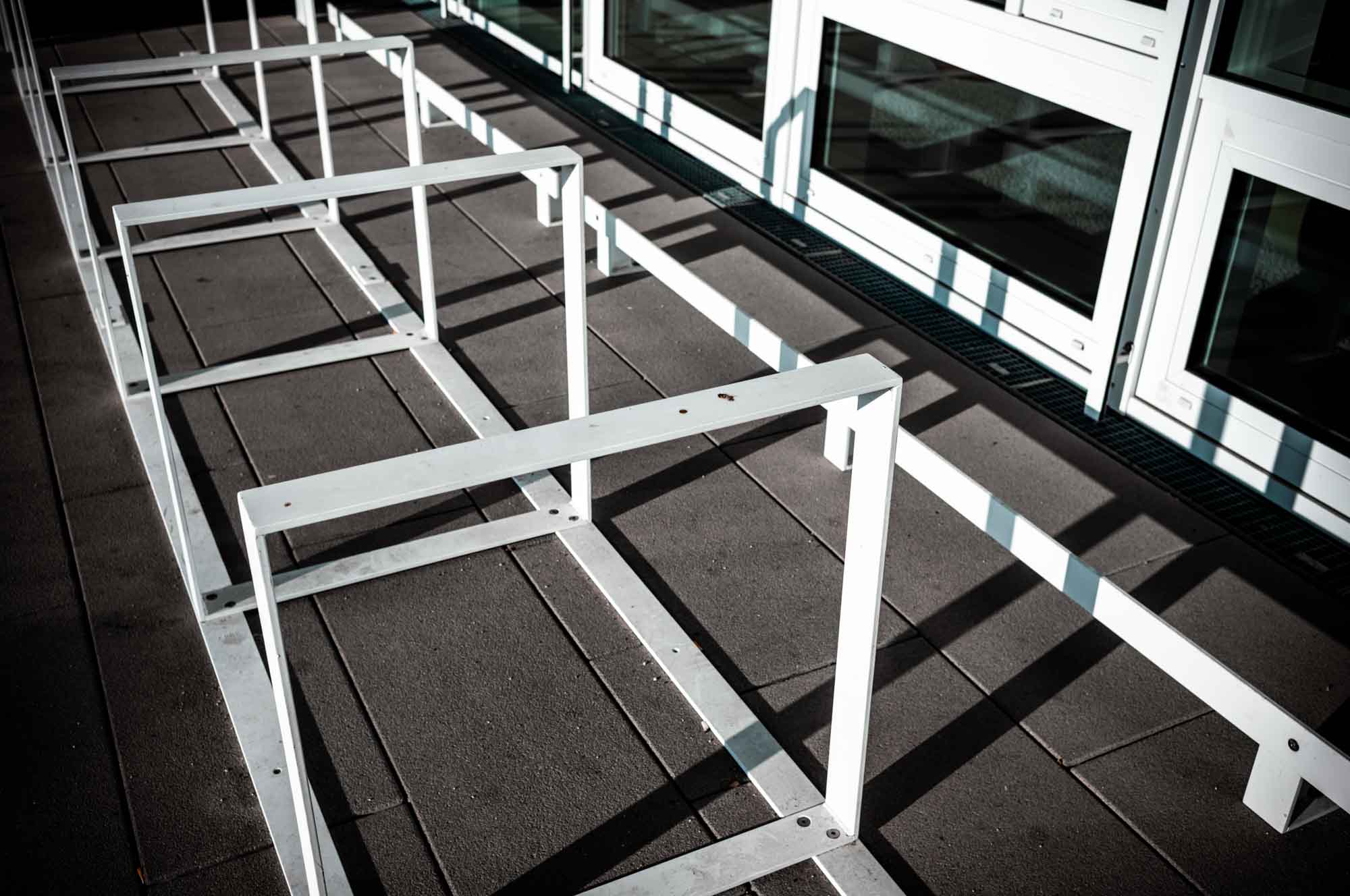 Modern white metal bike rack casting shadows on a concrete surface by glass windows.