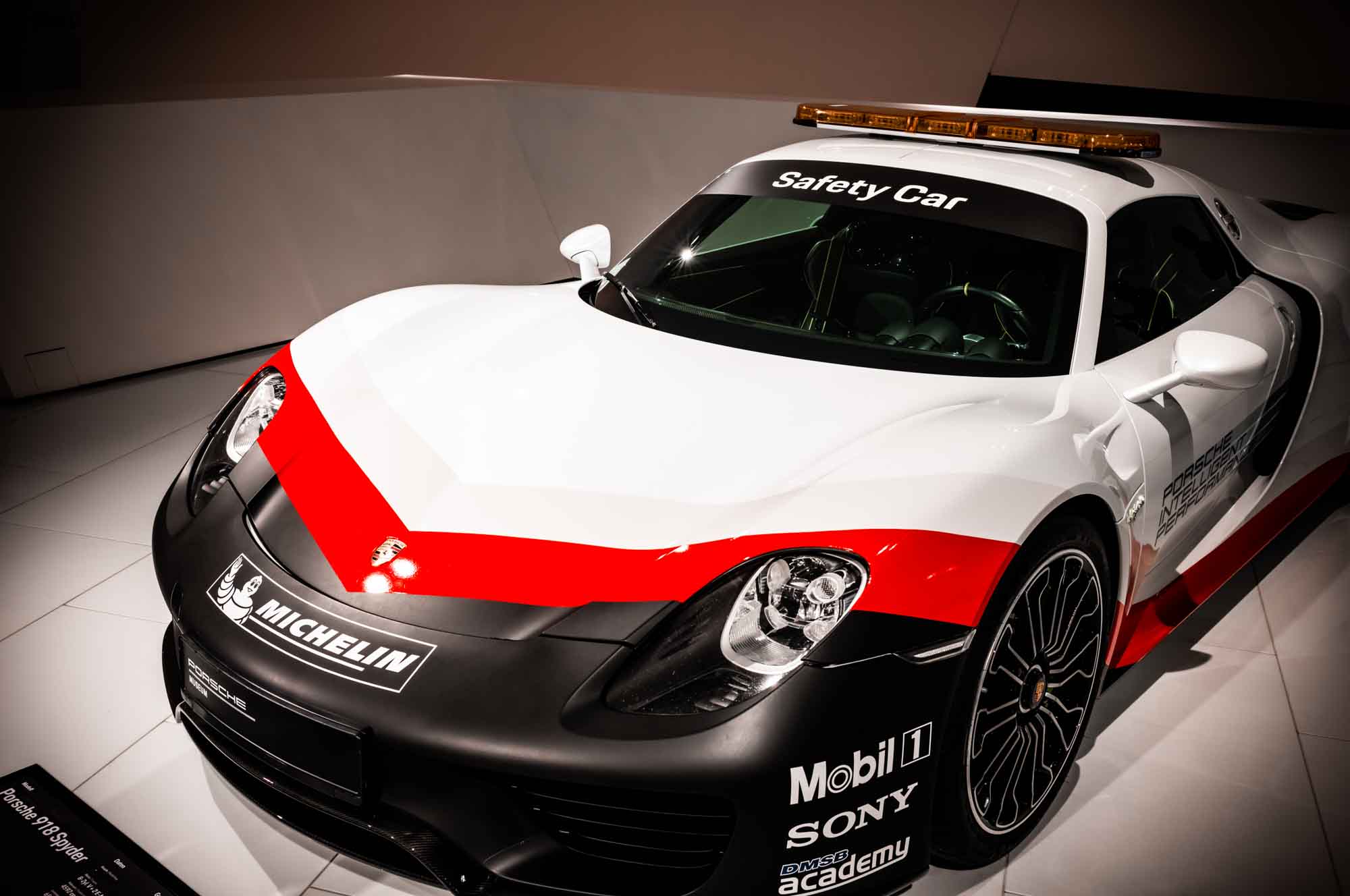 White and red Porsche 918 hypercar labeled Safety Car displayed in a showroom with sponsor decals.