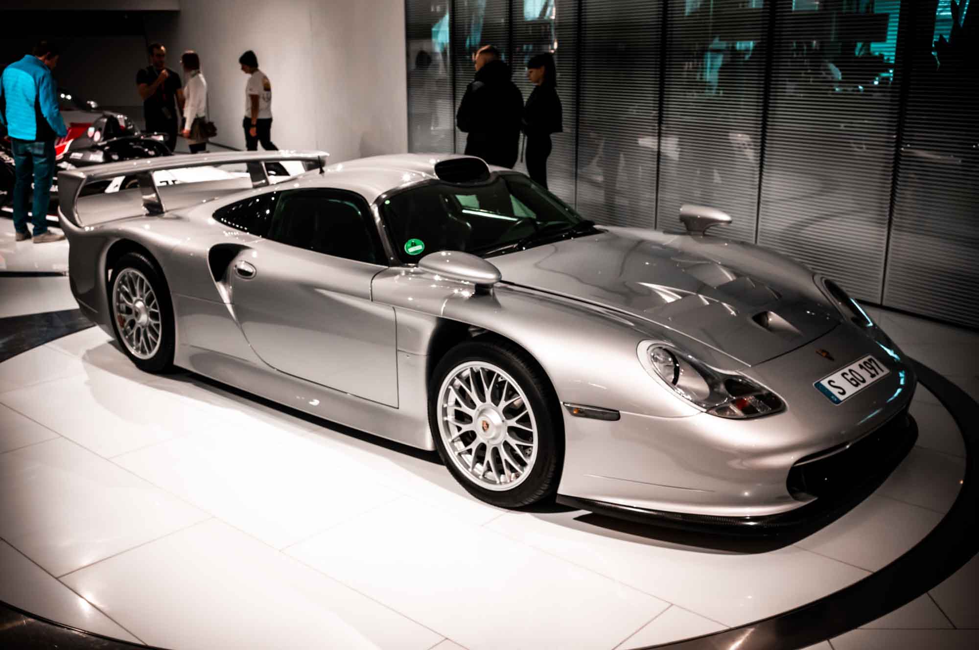Silver sports car displayed in a modern showroom with people observing in the background.
