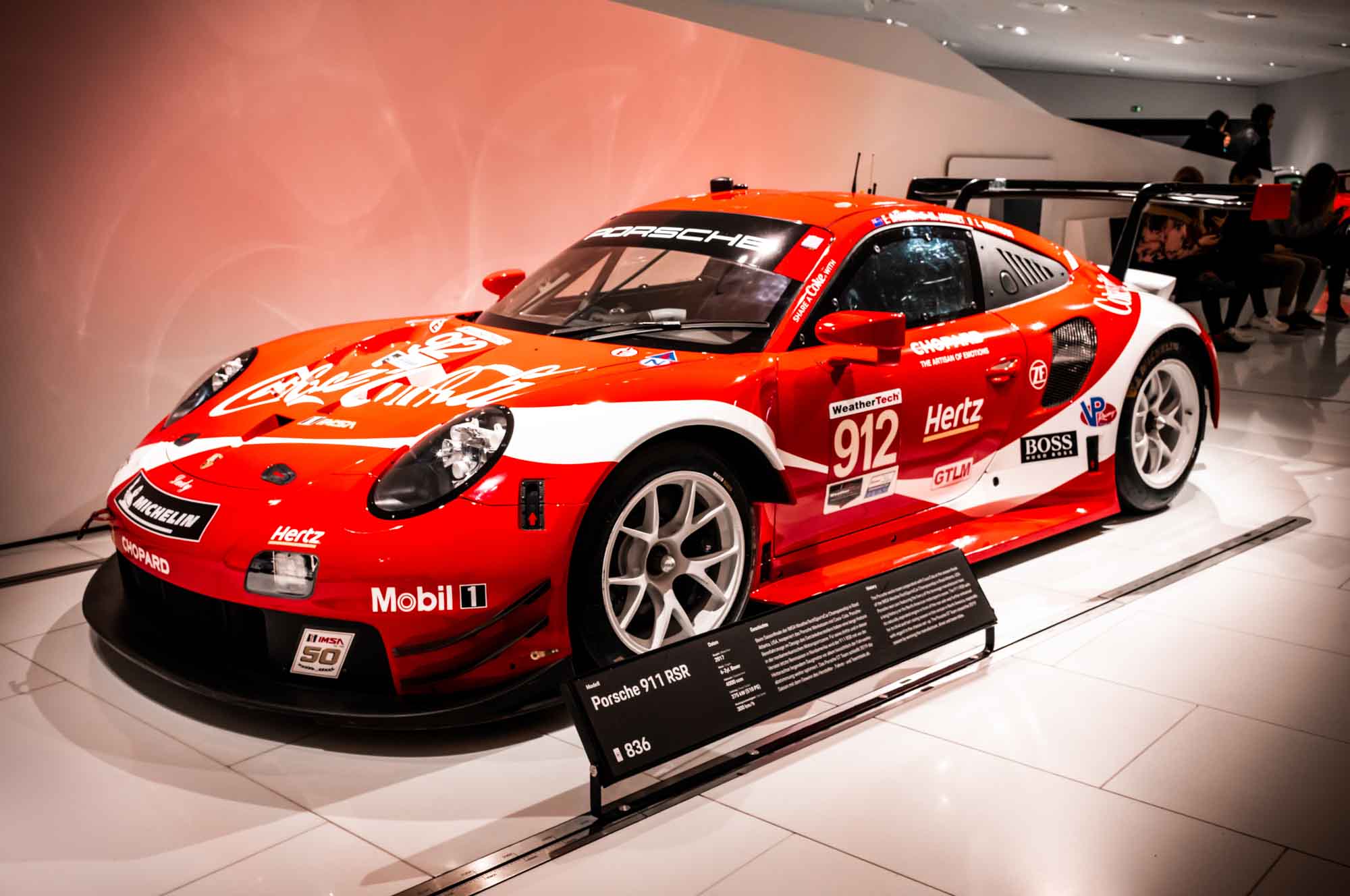 Red Porsche 911 RSR race car displayed in a showroom, featuring sponsor logos and a detailed specification plaque.