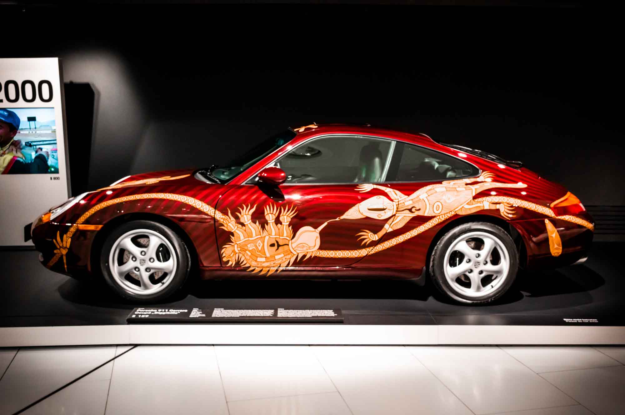 Sports car with intricate orange lizard artwork on a display platform in a dimly lit exhibition space.