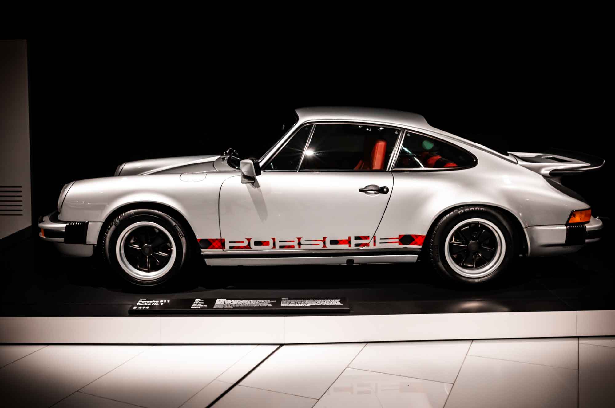 Silver classic sports car on display, side view with red accents and sleek design, showcased in a modern showroom setting.