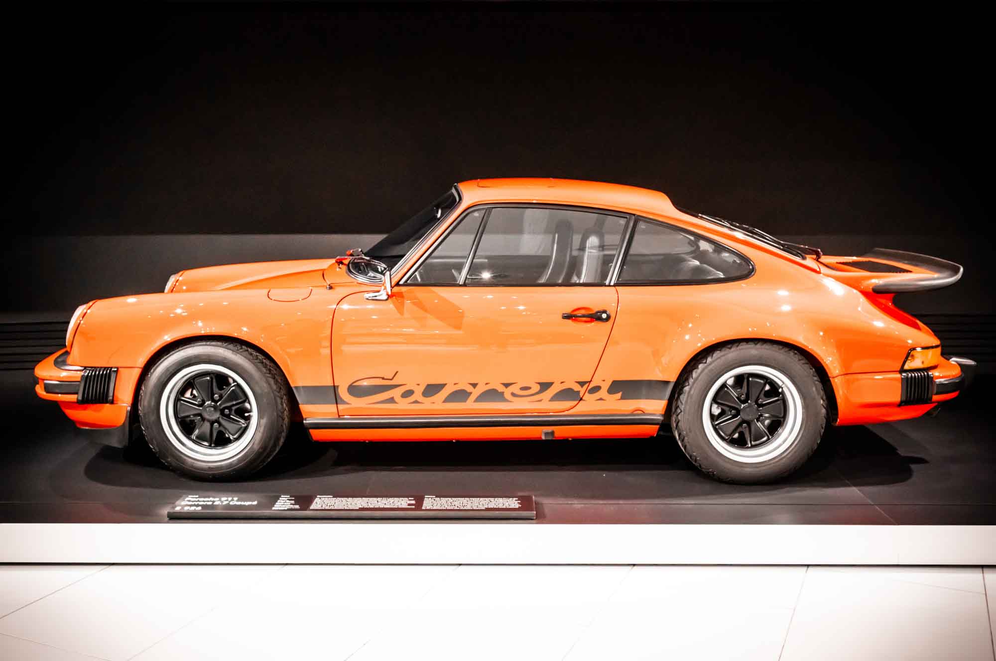 Orange vintage sports car parked in a showroom, side profile view.