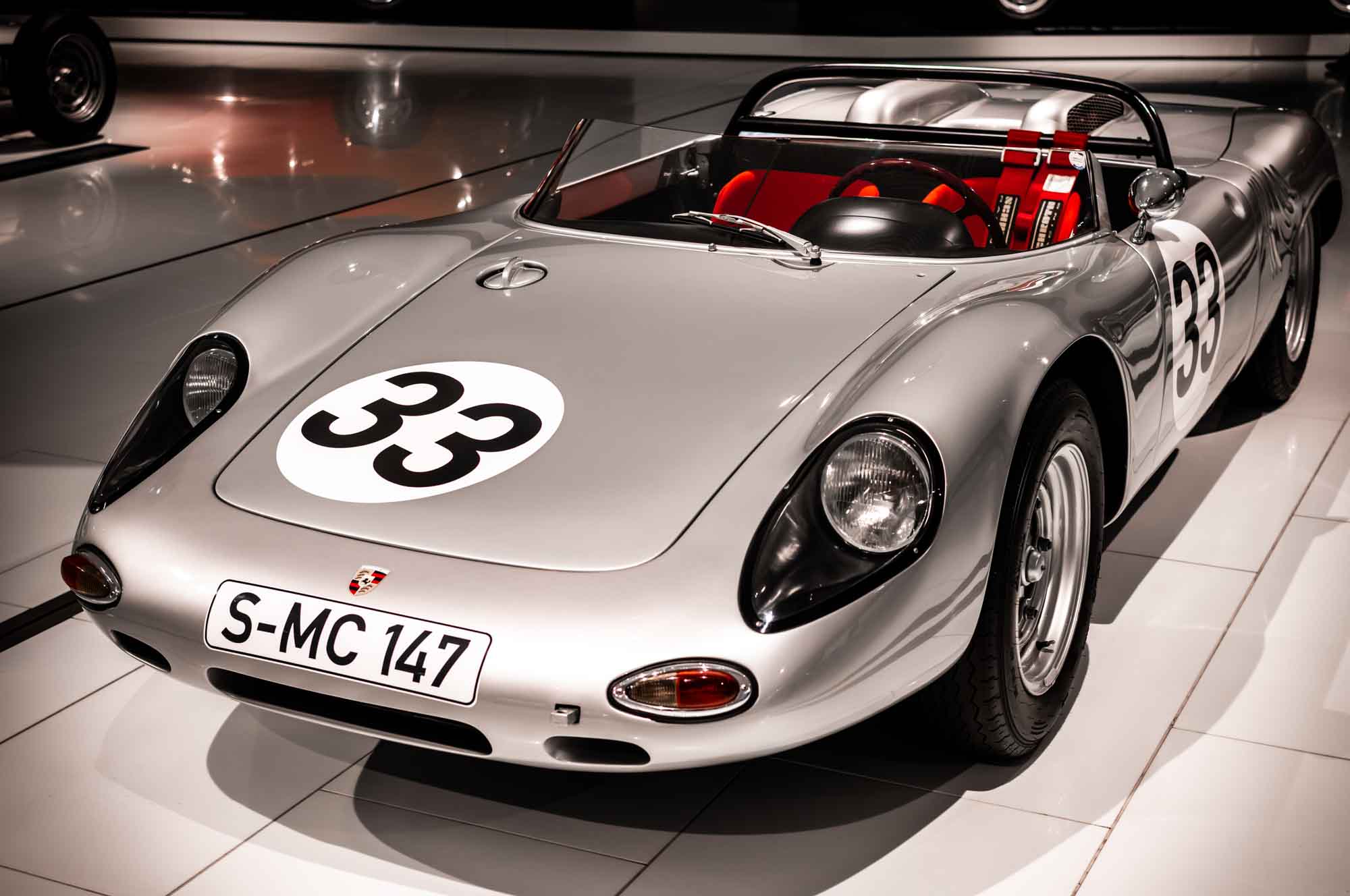 Silver vintage racing car with number 33 displayed on the hood, parked in a showroom.