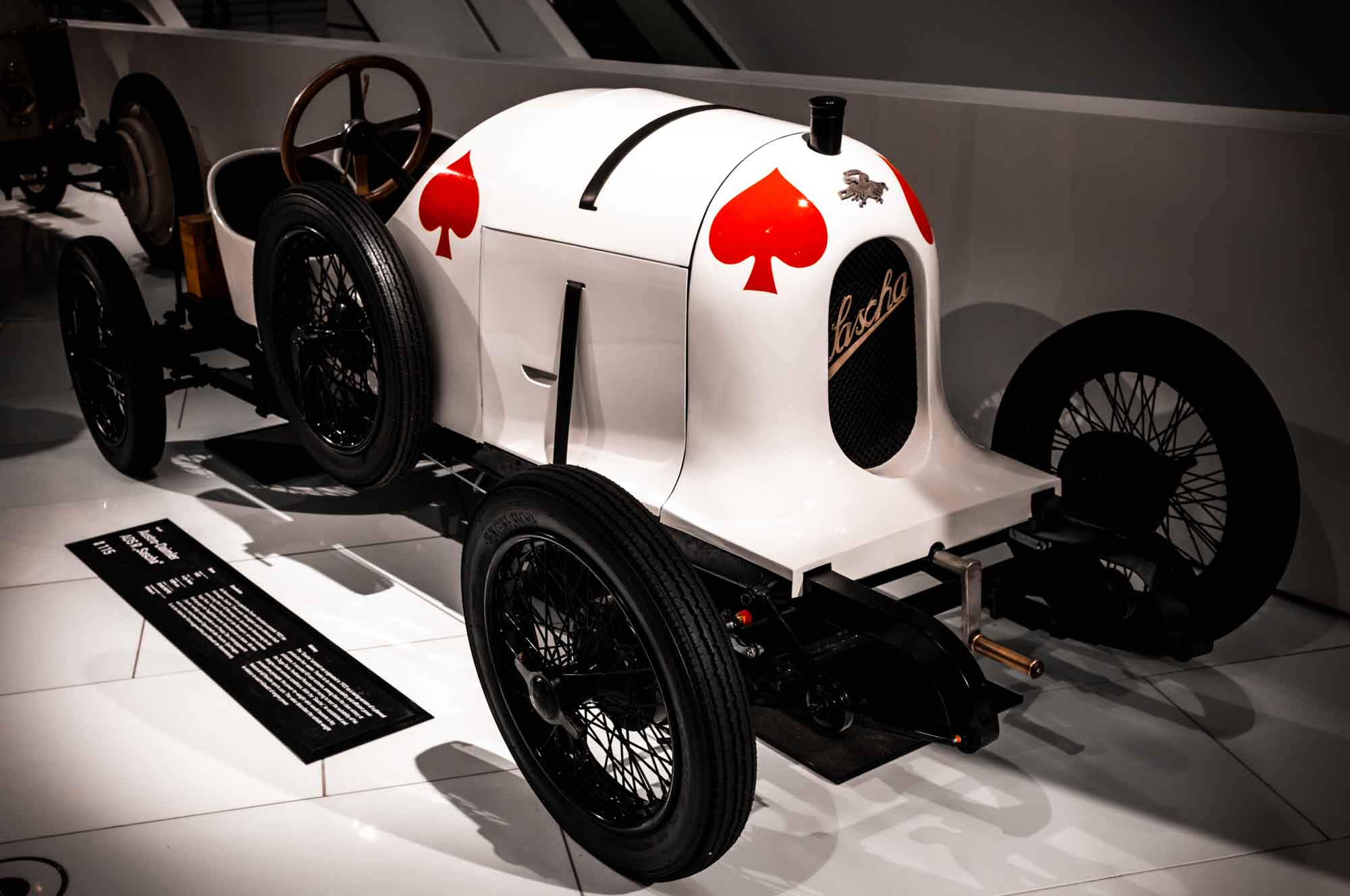 Vintage white race car with red spades, wooden steering wheel, and spoked wheels on display in a museum.