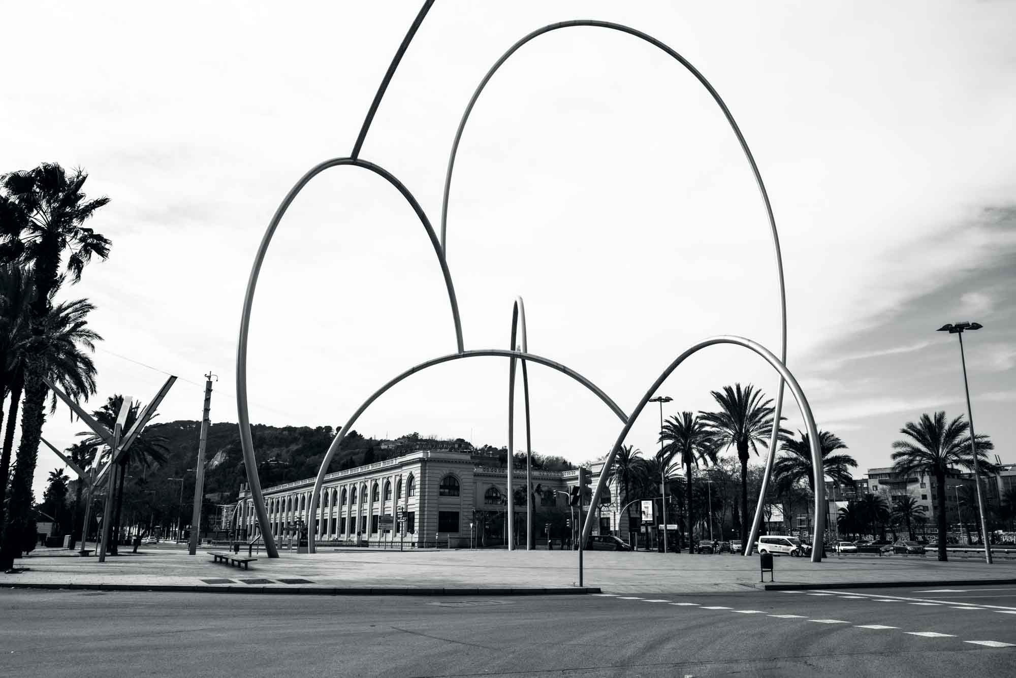 Modern urban sculpture with arches and palm trees in a cityscape, showcasing architectural design.