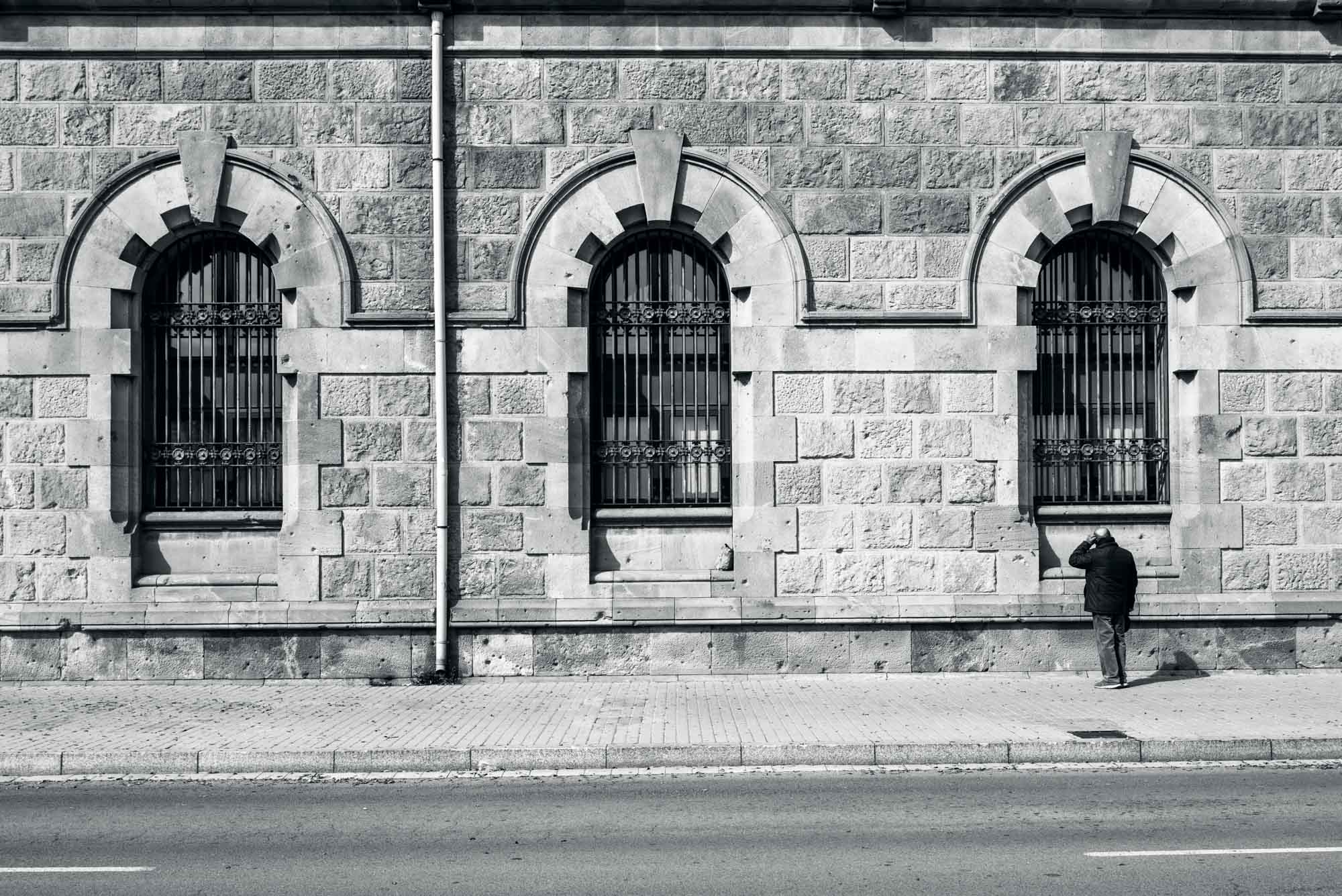Black and white photo of a person walking past a historic stone building with arched windows.