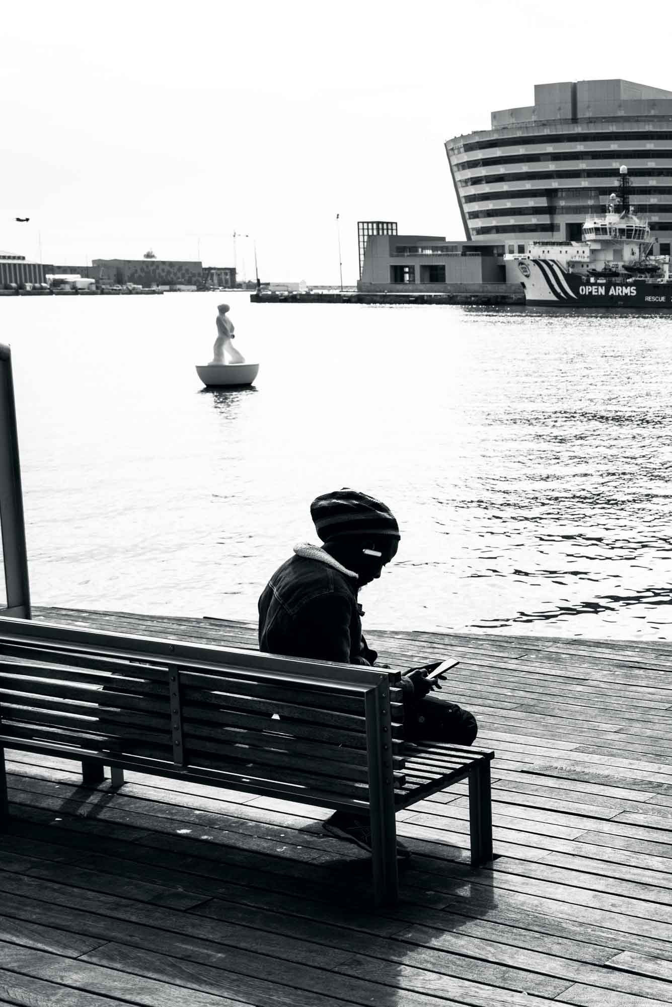 Silhouette of a person on a bench by a waterfront, with a ship and a floating sculpture in the background.