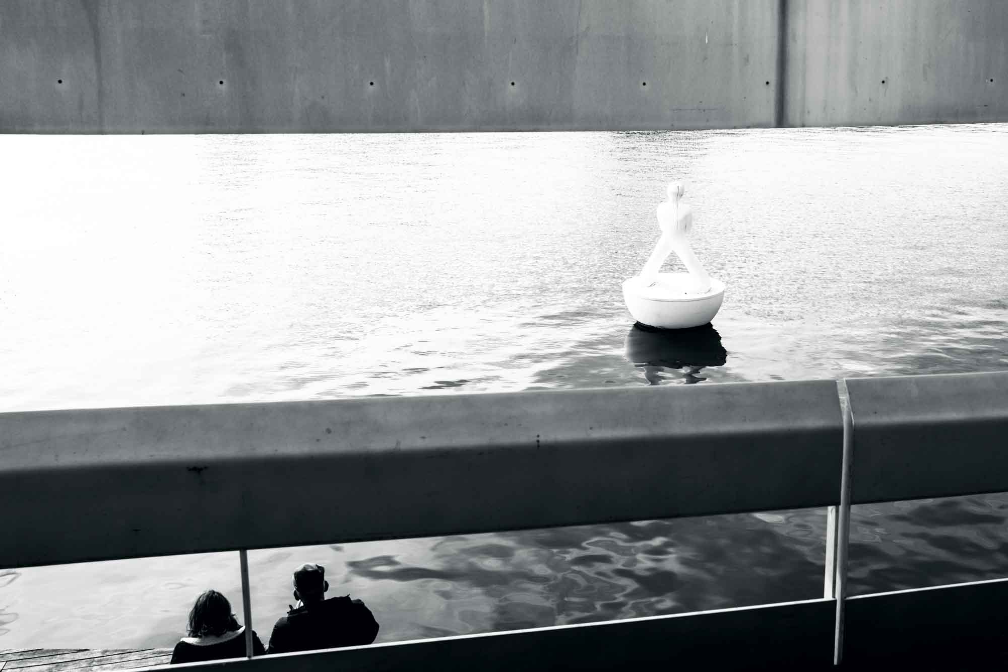 Black and white photo of two people sitting by a riverside, near a water sculpture under a modern bridge.