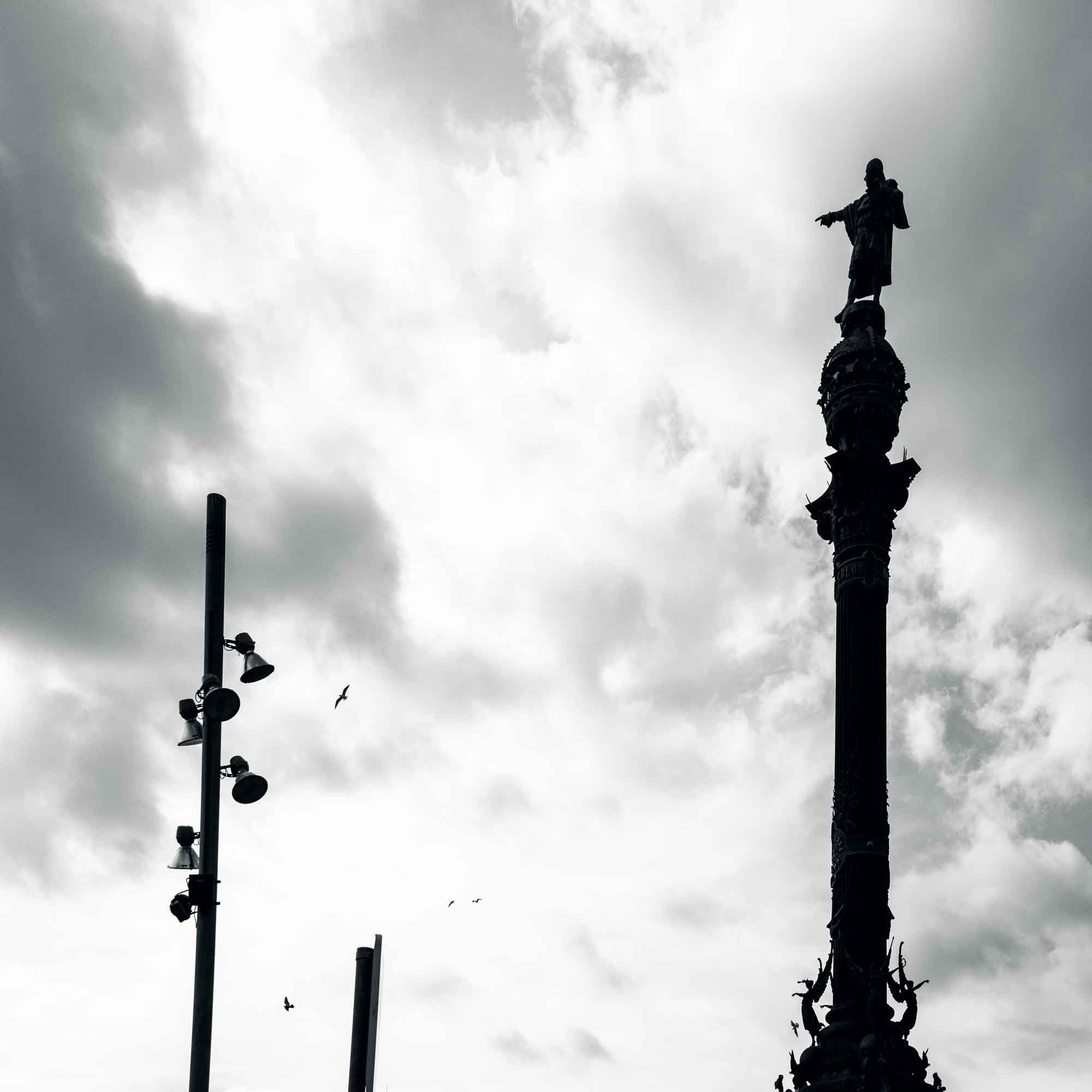 Silhouette of a monument and streetlights against a cloudy sky in black and white.