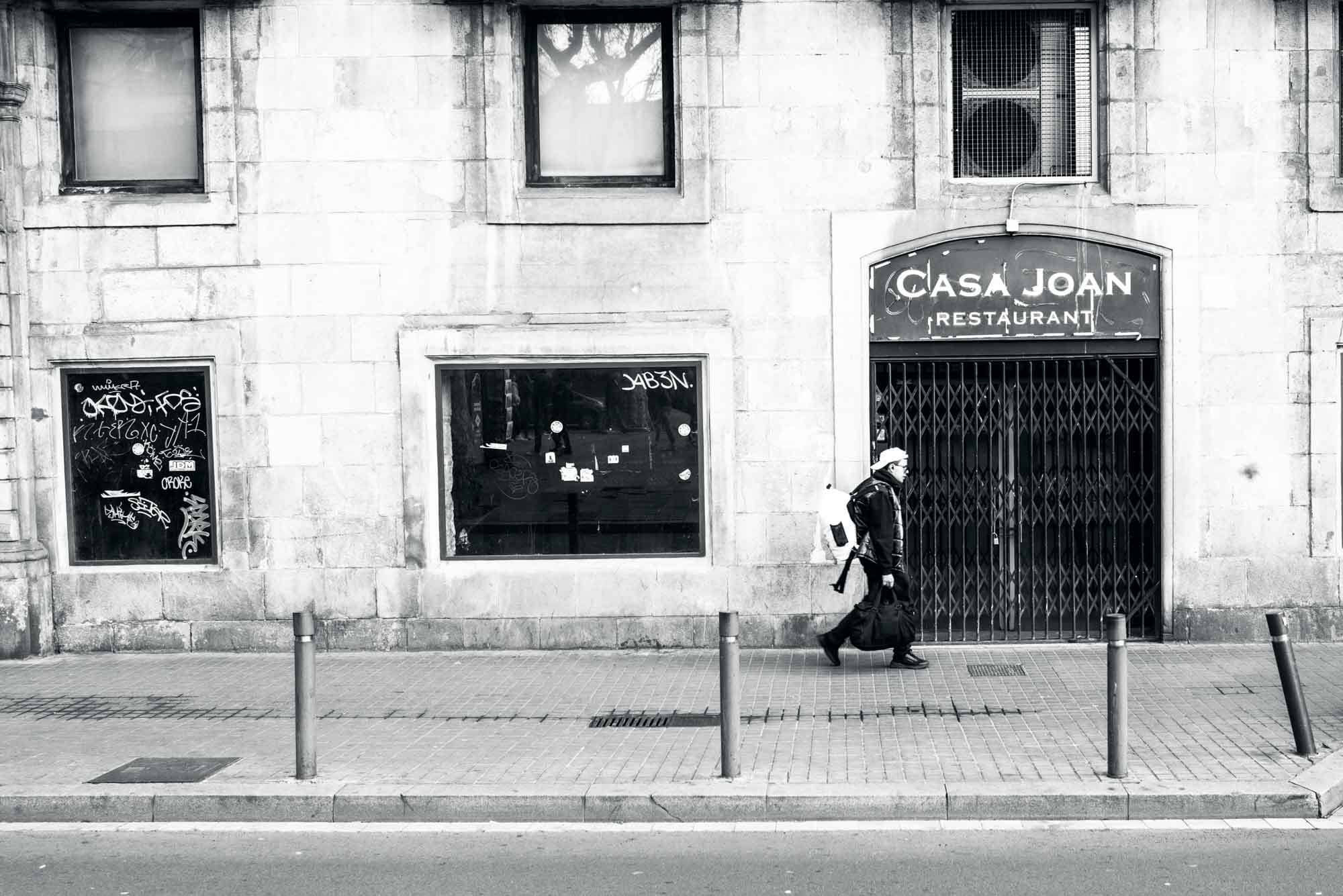 Person walking past closed Casa Joan restaurant with a backpack, grayscale urban street scene.