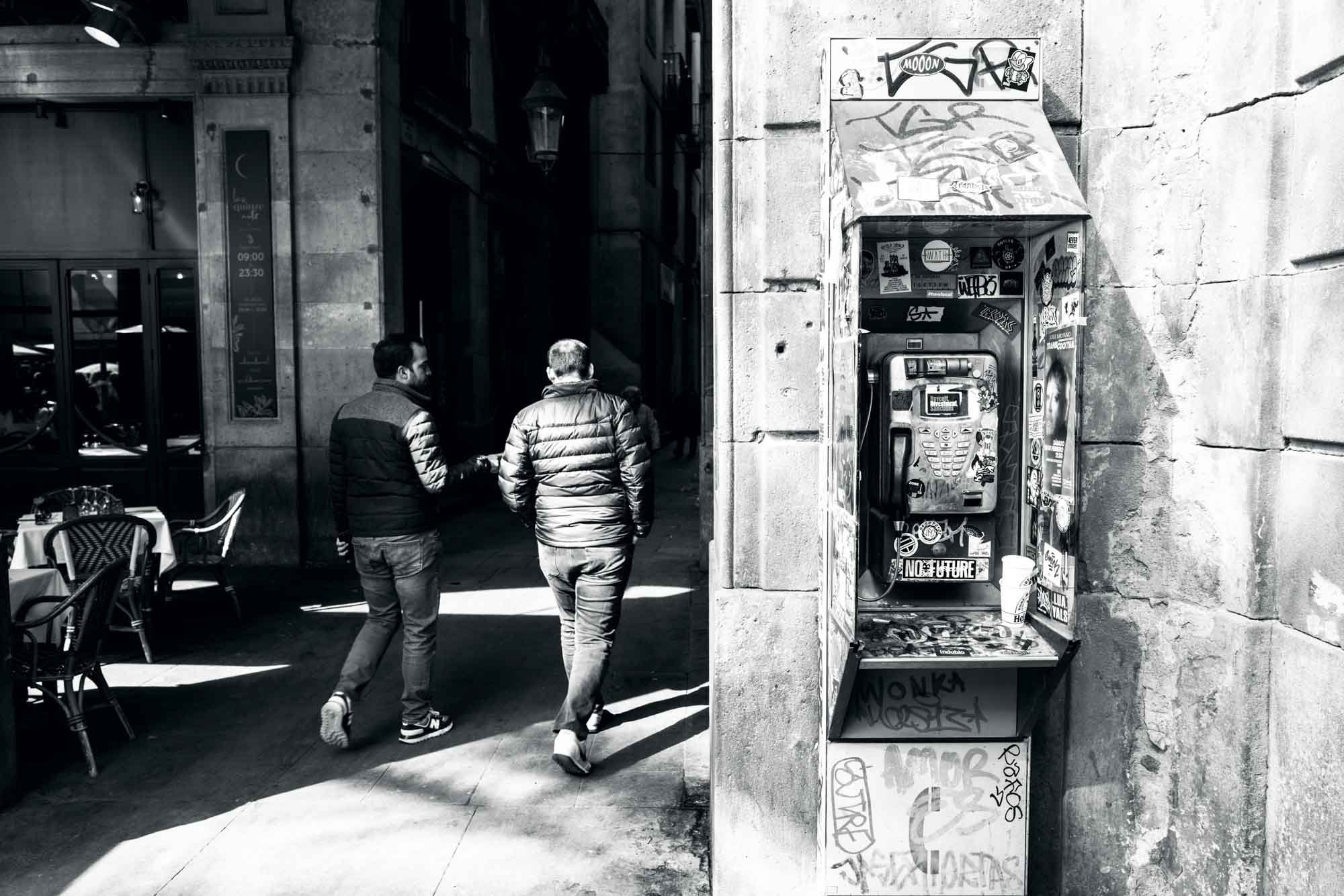 Urban street scene with two men walking, passing a graffitied payphone on a sunny day.