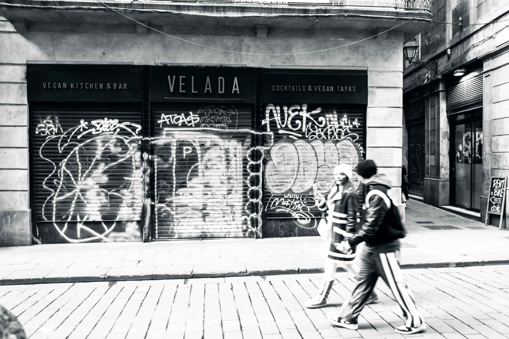 Street view of pedestrians passing a closed vegan bar with graffiti-covered shutters in black and white.