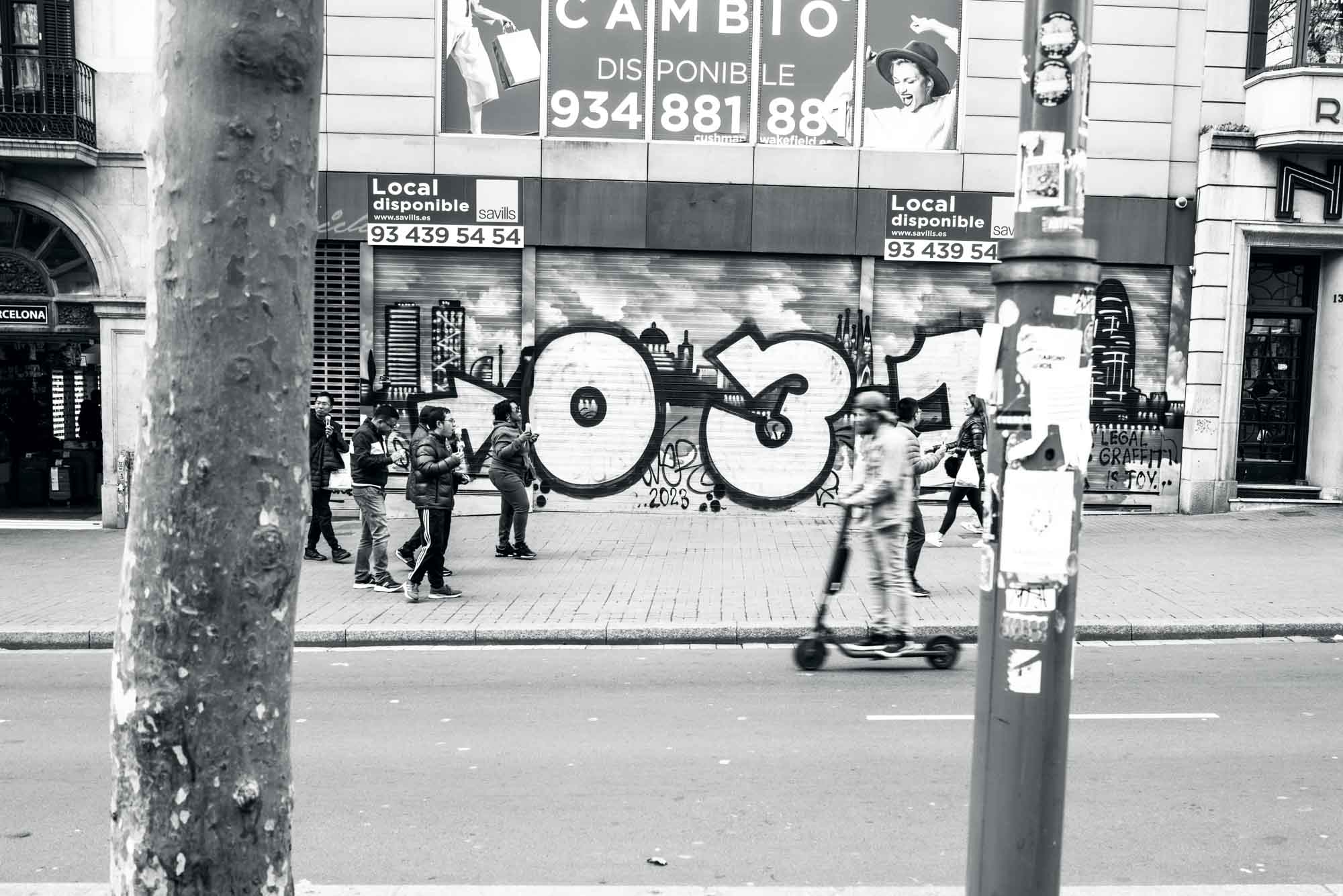 Street scene with people walking and riding a scooter in front of graffiti-covered building in a city.