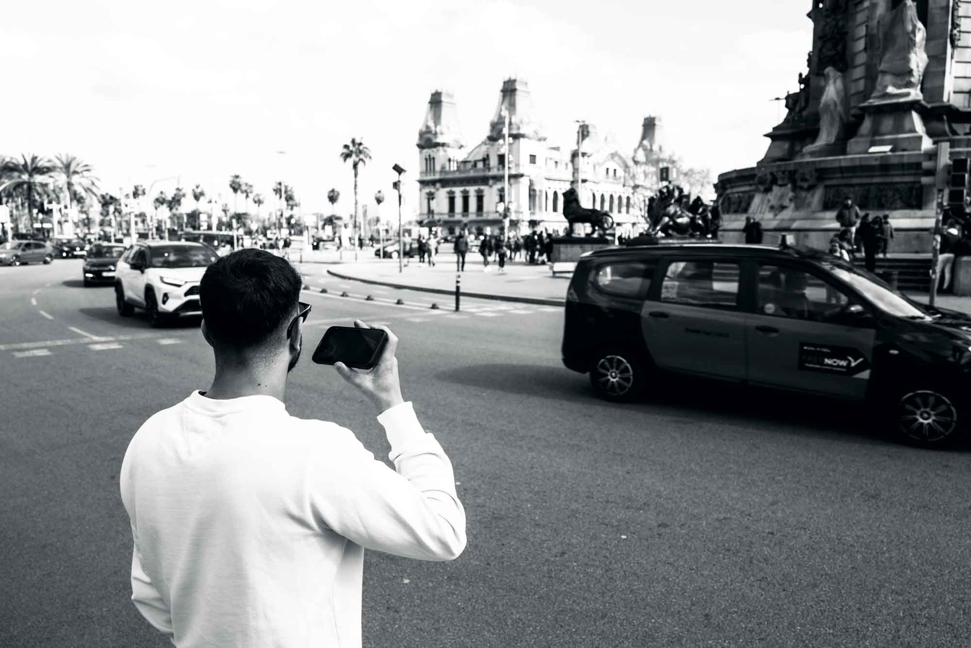 Man in white shirt captures busy city street with phone camera, featuring cars and historic buildings in the background.