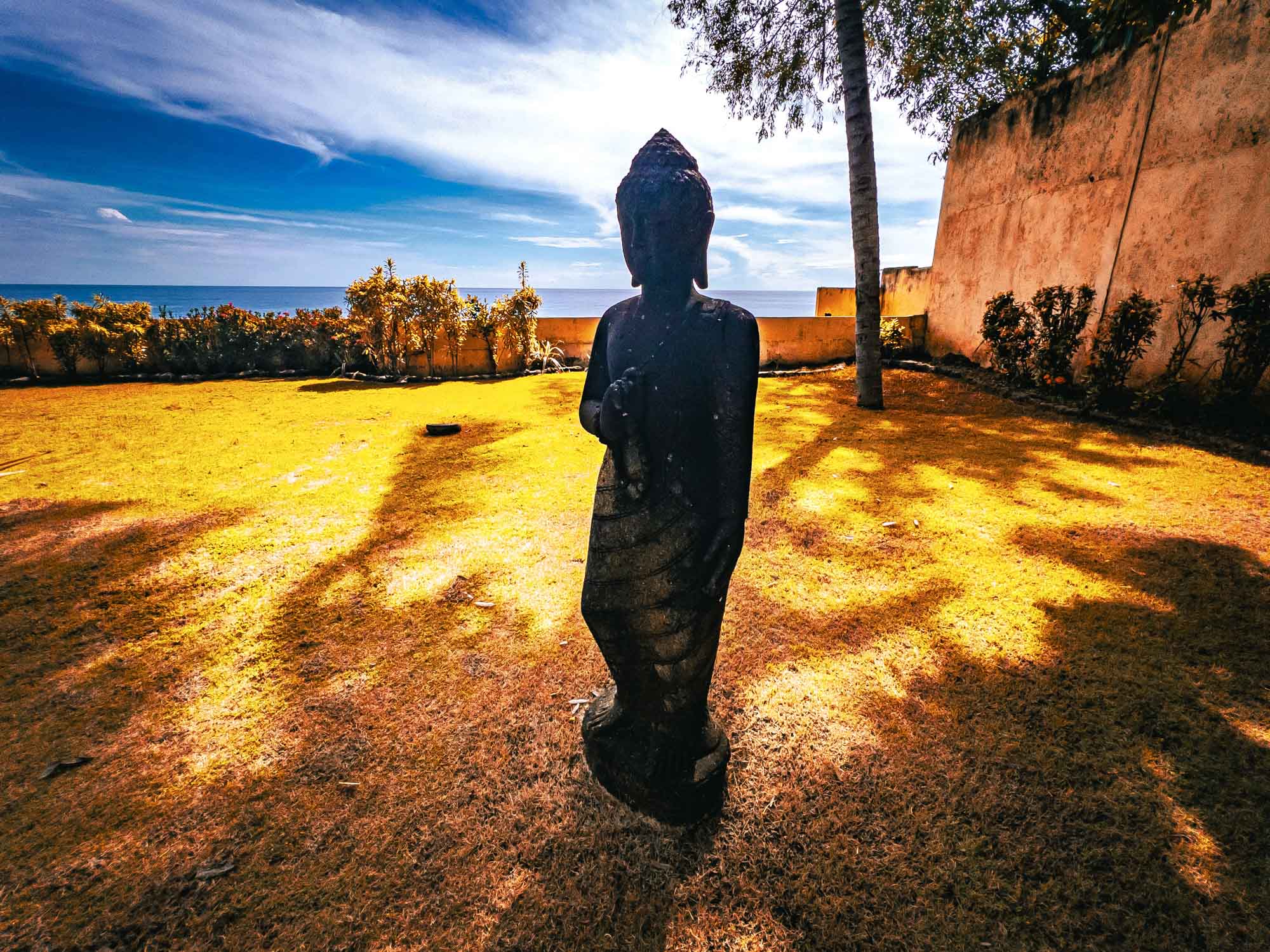 Statue in a sunlit garden near the sea with vibrant yellow grass and a clear blue sky.