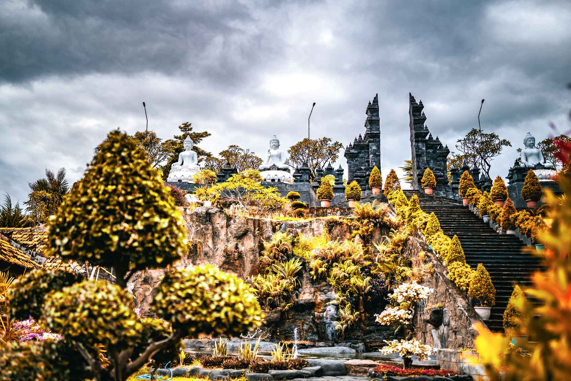 Temple surrounded by lush greenery and statues under a dramatic sky.
