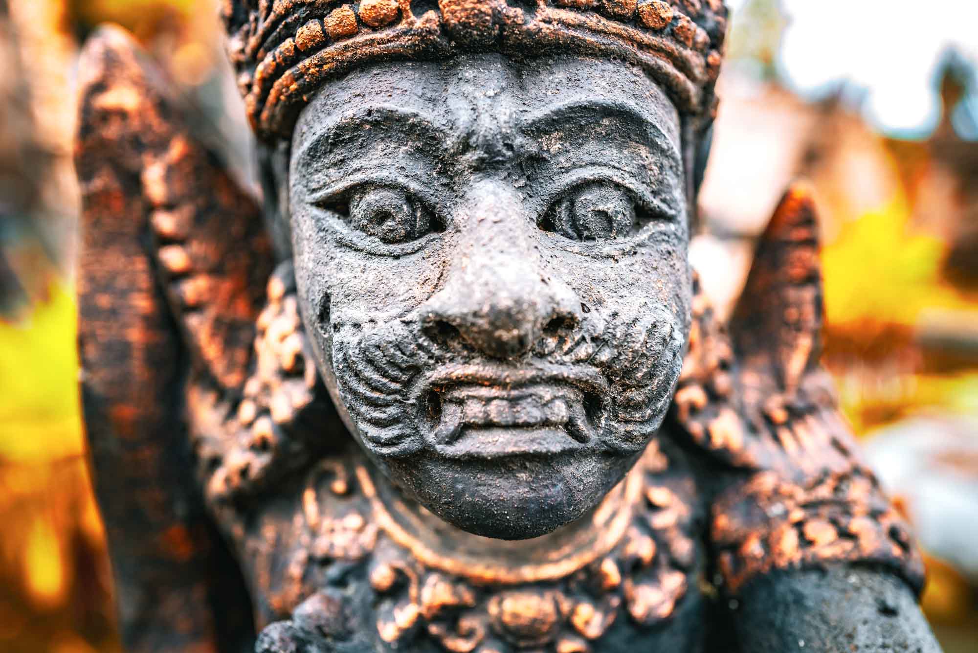 Close-up of an intricate stone statue face, featuring detailed carvings and vibrant autumn colors in the background.