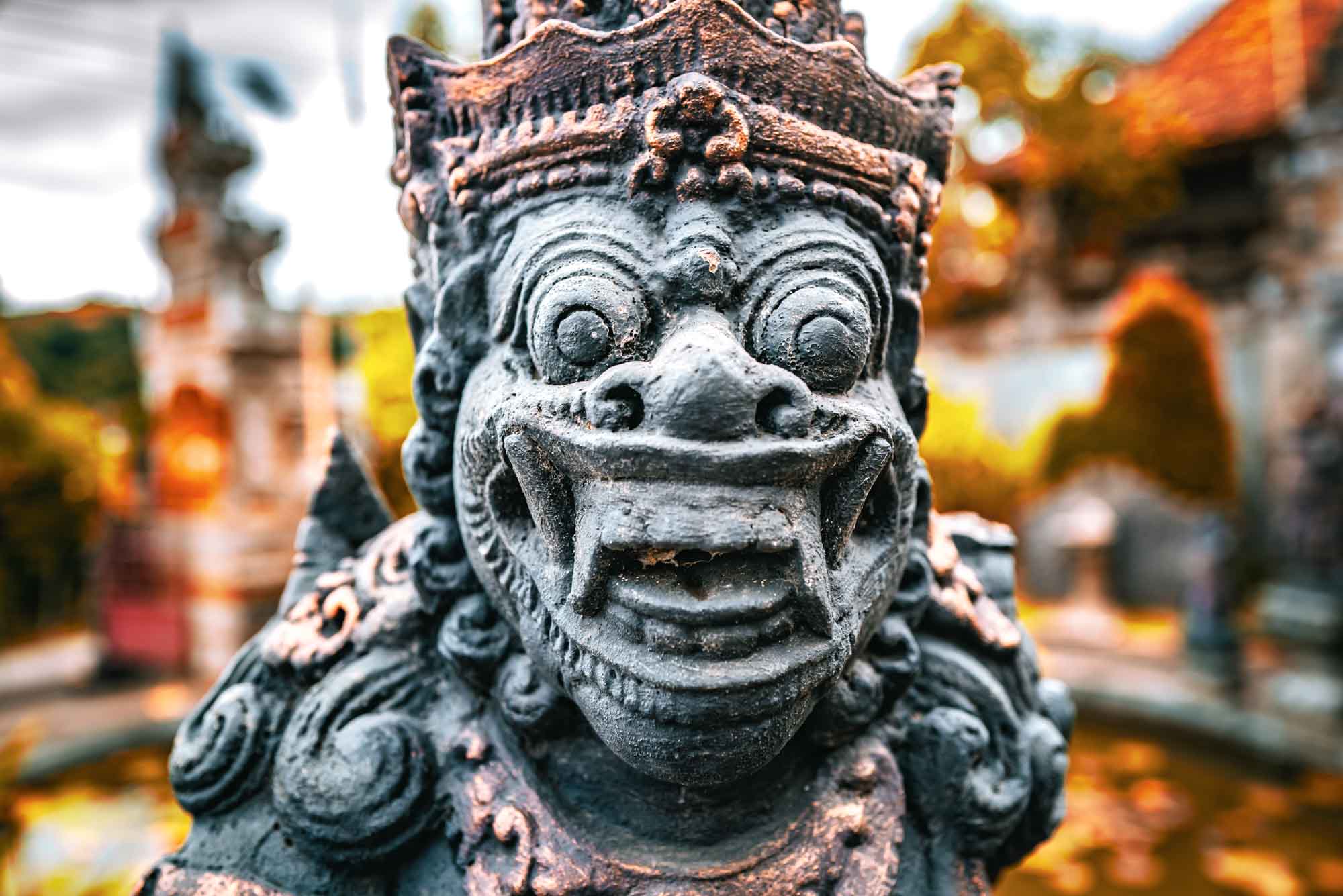 Ancient Balinese stone statue with intricate carvings in an outdoor setting, surrounded by blurred greenery and temple structures.
