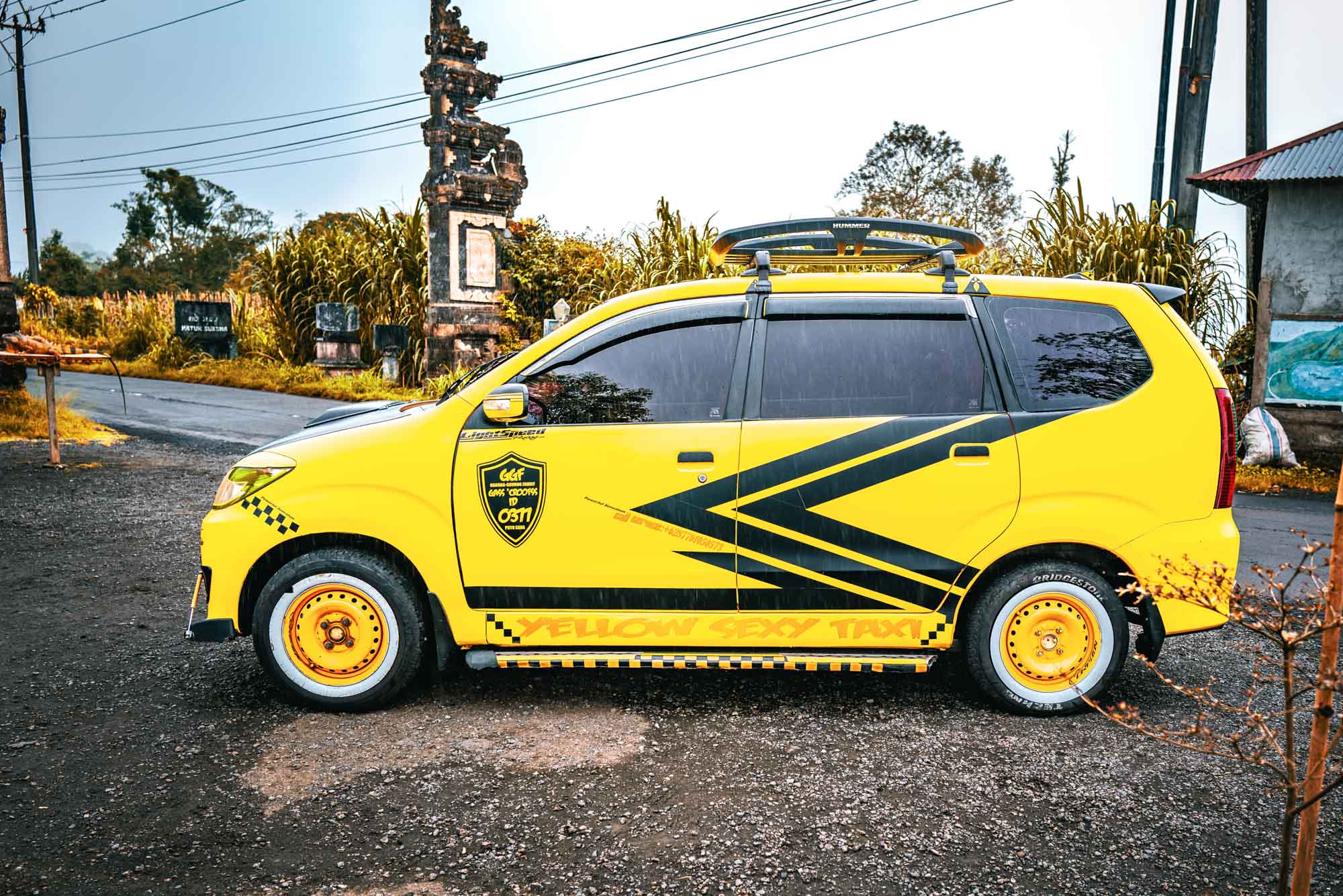 Bright yellow taxi with bold black stripes parked on a rural road, surrounded by greenery and traditional architecture.