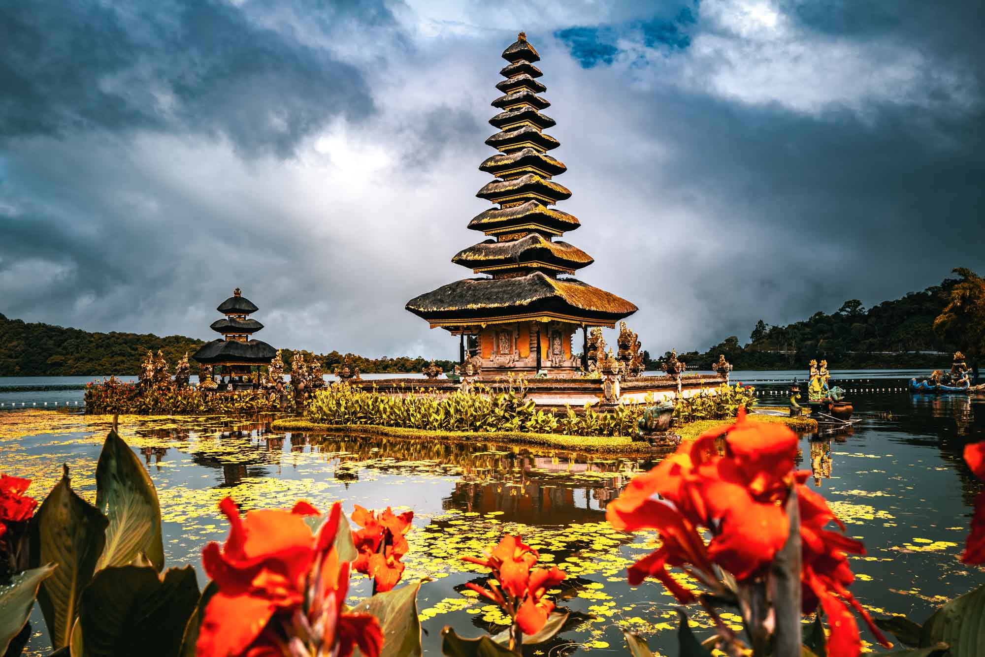 Balinese temple on a lake surrounded by vibrant red flowers and dramatic clouds in the background.