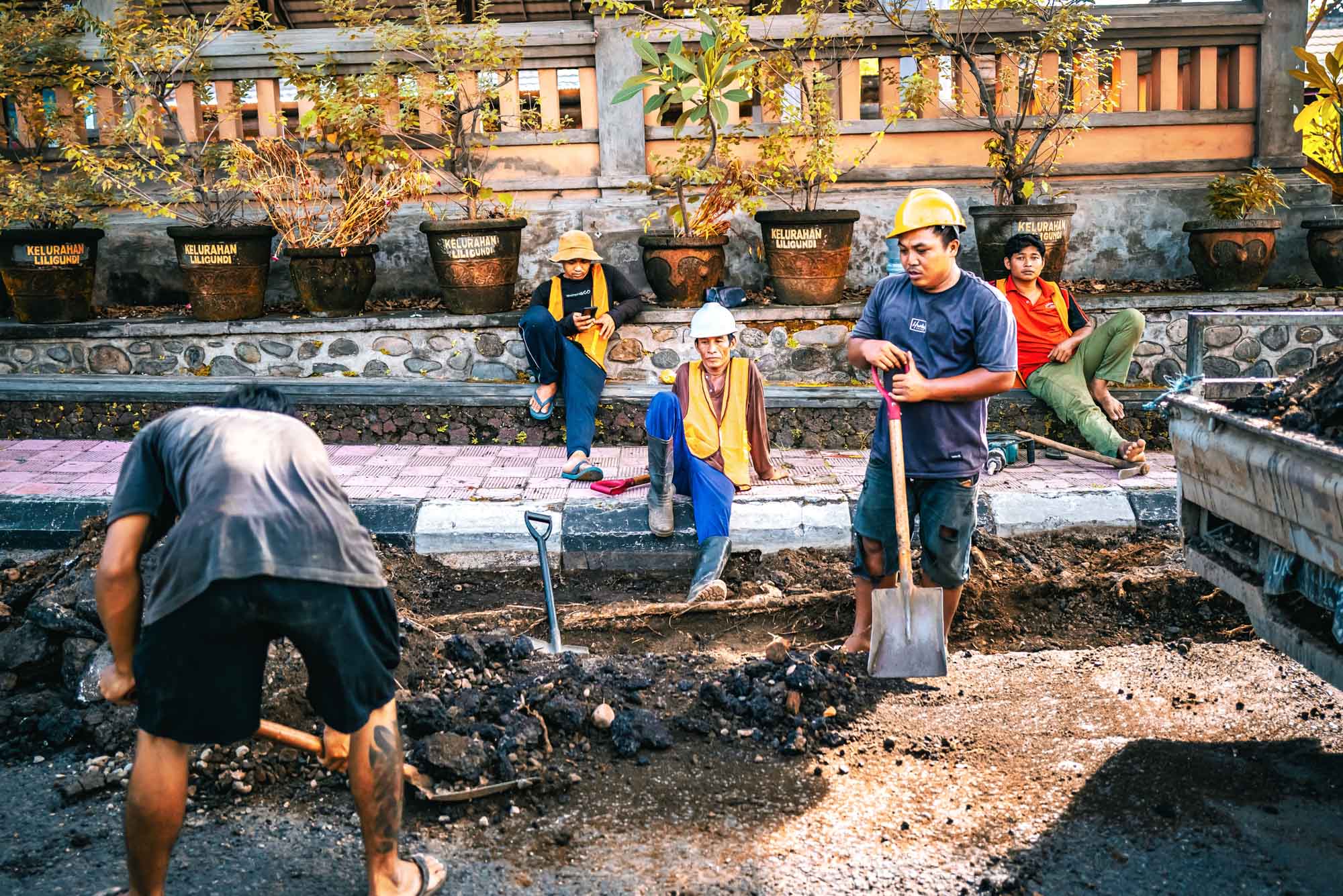 Construction workers repairing road while others take a break. Shovels and debris are visible. Urban setting with plants.