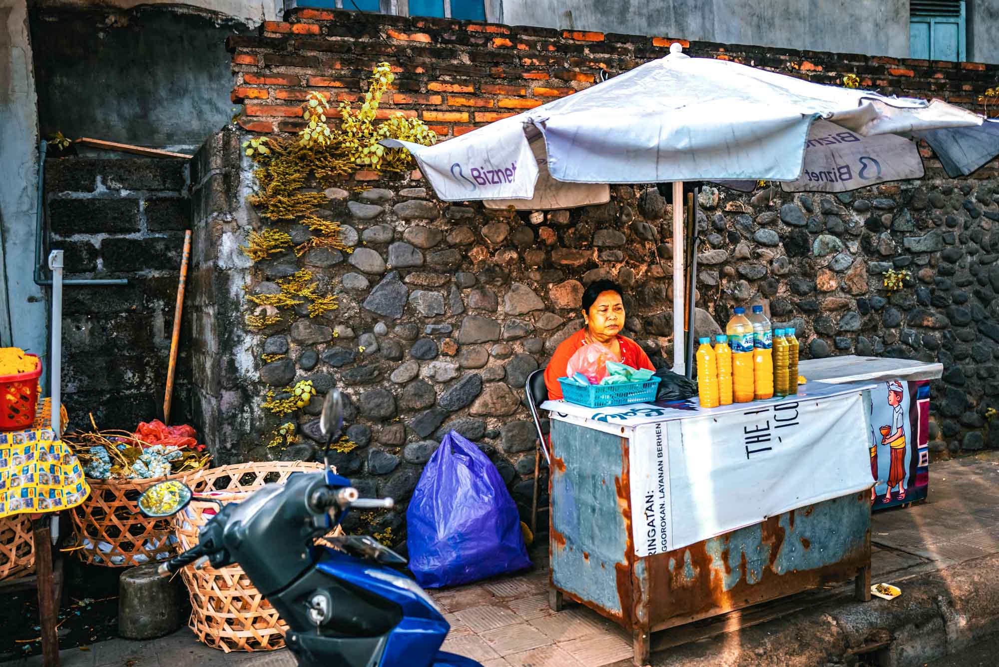 Street vendor under umbrella selling bottled yellow beverages in front of a stone wall, next to a parked scooter.