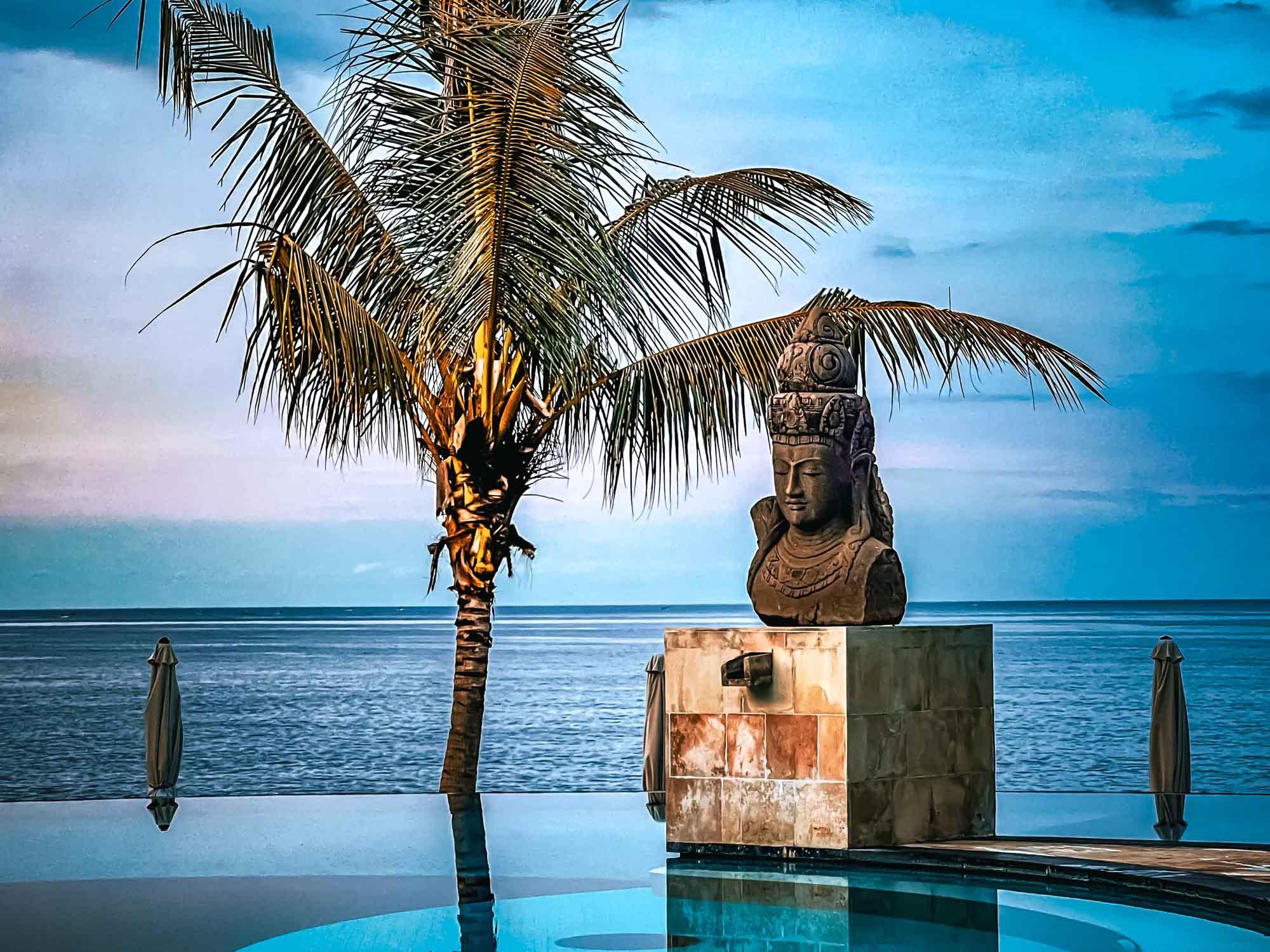 Tropical scene with palm tree and statue by a serene ocean under a blue sky, ideal for a peaceful getaway.