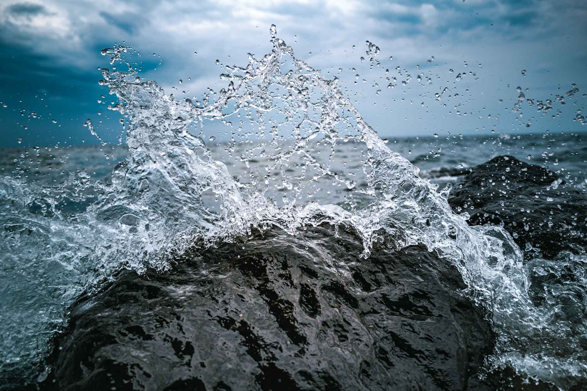Dynamic ocean wave splashing over dark rocks under a cloudy sky.