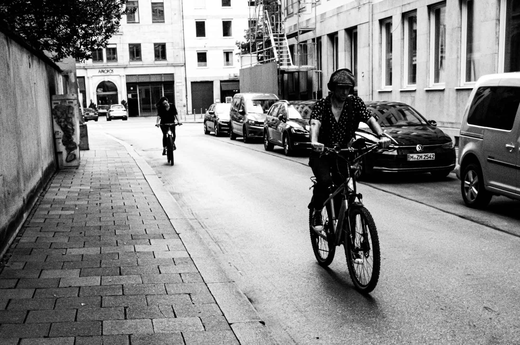 Two cyclists ride down an urban street, lined with parked cars and buildings, captured in black and white.