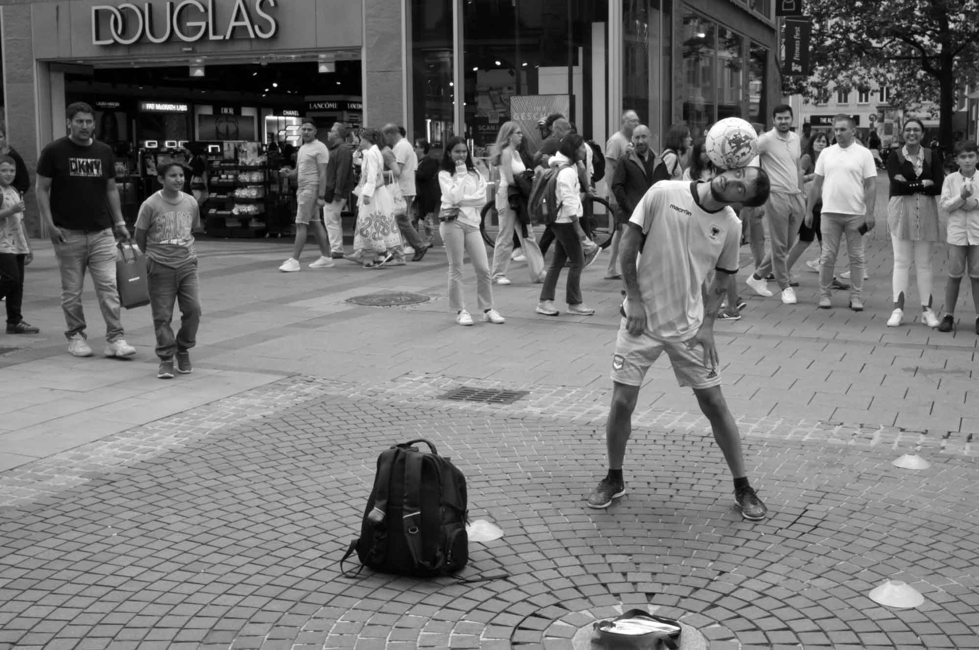 Street performer balances soccer ball on head in busy shopping area, attracting a crowd of onlookers.
