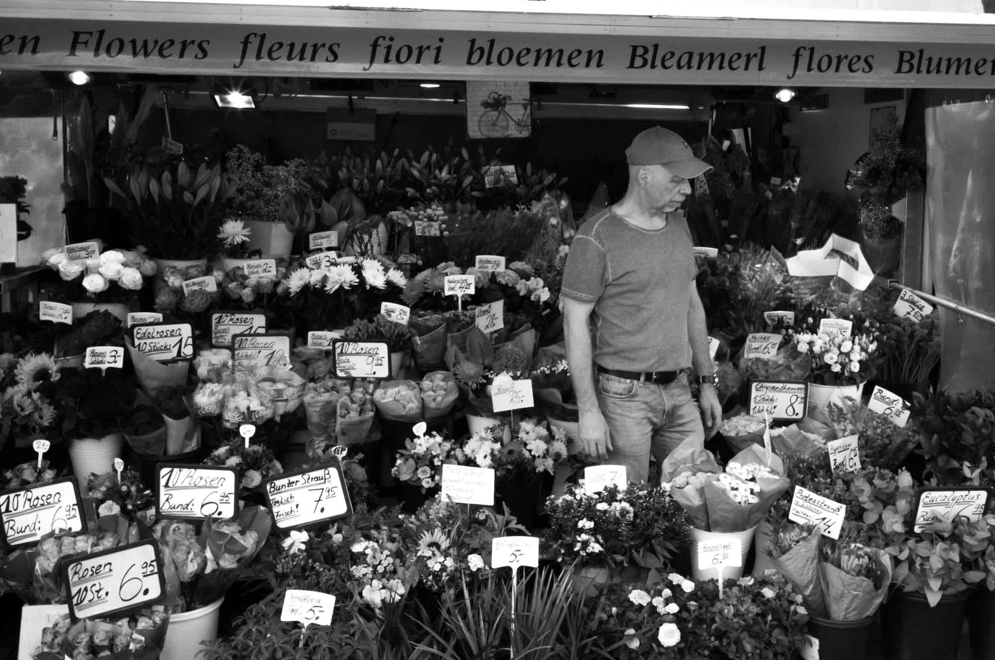 Man in front of vibrant flower market stall with various blooms and price tags in multiple languages.