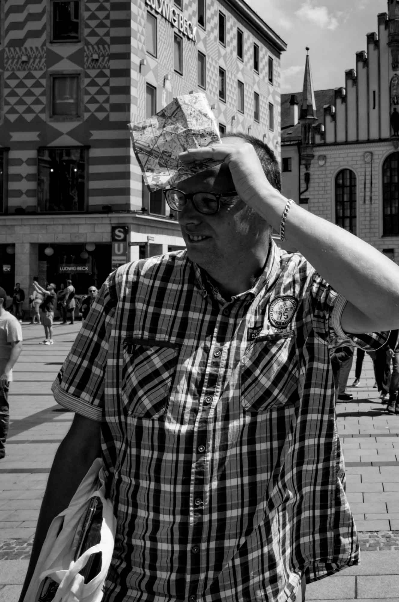 Man holding a city map in a bustling plaza, wearing glasses and plaid shirt, architecture in the background.