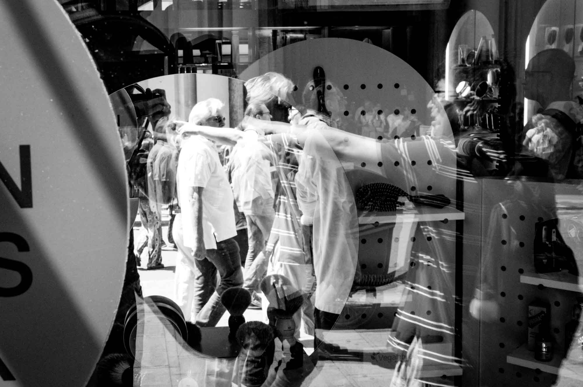 Black and white reflection of urban street scene in storefront window, capturing people walking and shop displays overlapping.