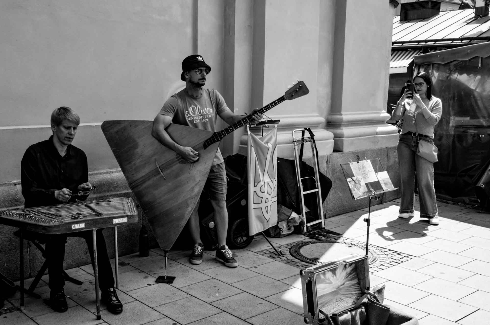 Street musicians with a large balalaika and cimbalom perform while a woman photographs them on a sunny day.