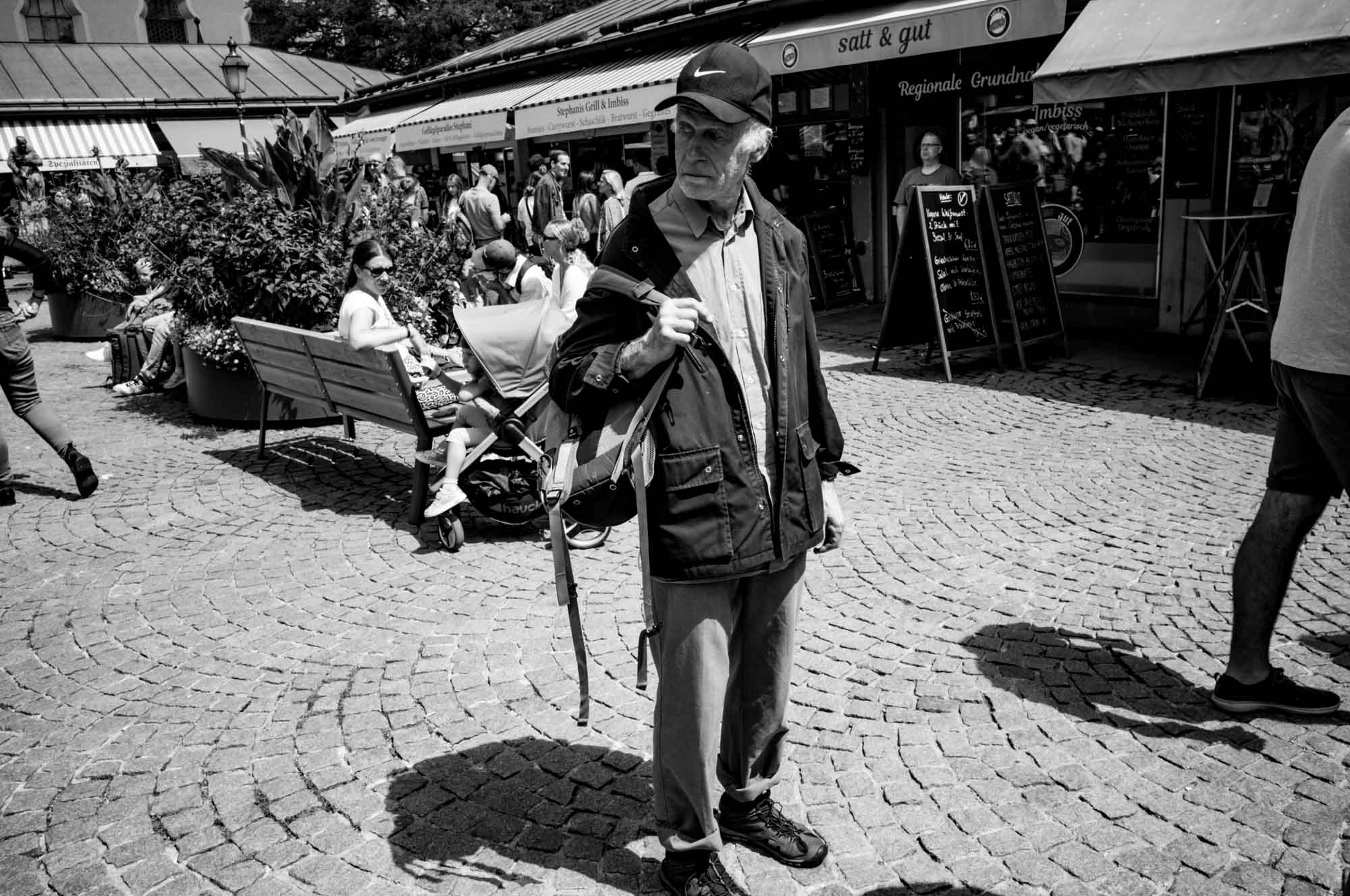 Black and white market scene with people walking and relaxing on benches under the sun.