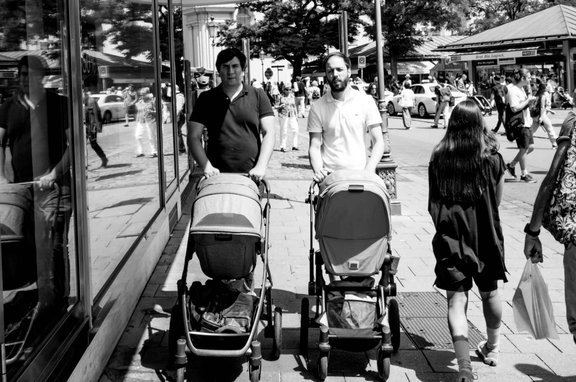 Two people walking with baby strollers on a busy city street reflection in shop window, black and white photo.