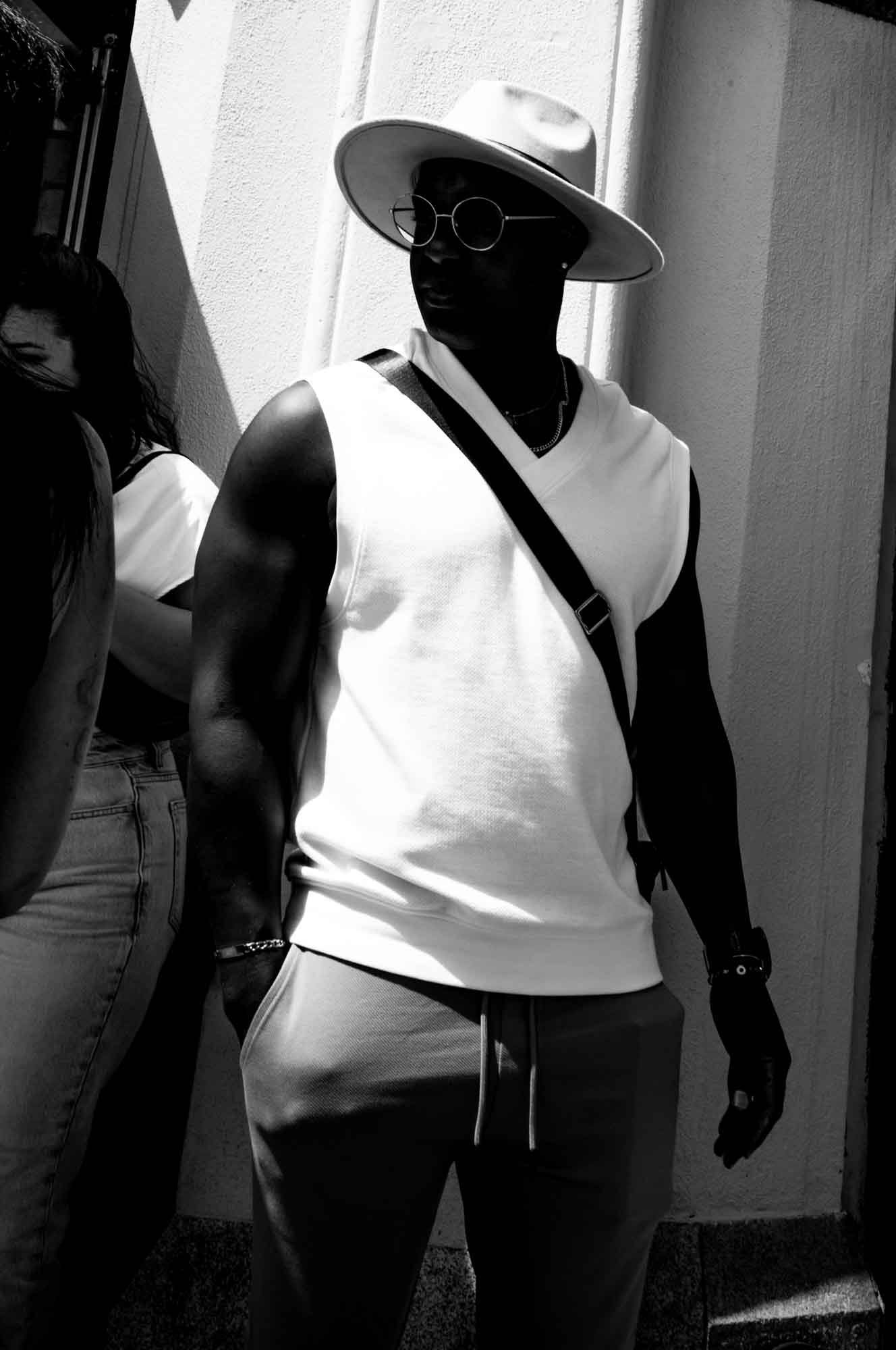 Fashionable man in a sun hat and sunglasses, wearing a sleeveless top, standing stylishly against a wall in black and white.