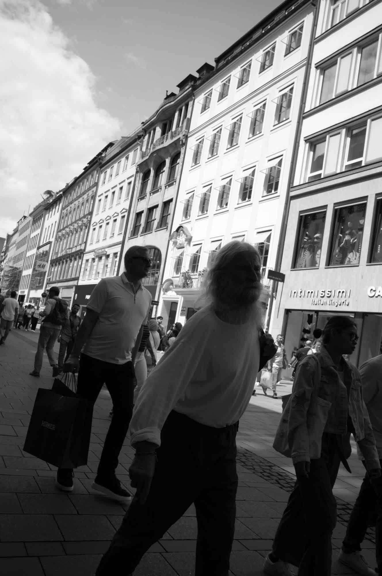 Black-and-white photo of people walking in front of European city buildings on a sunny day.