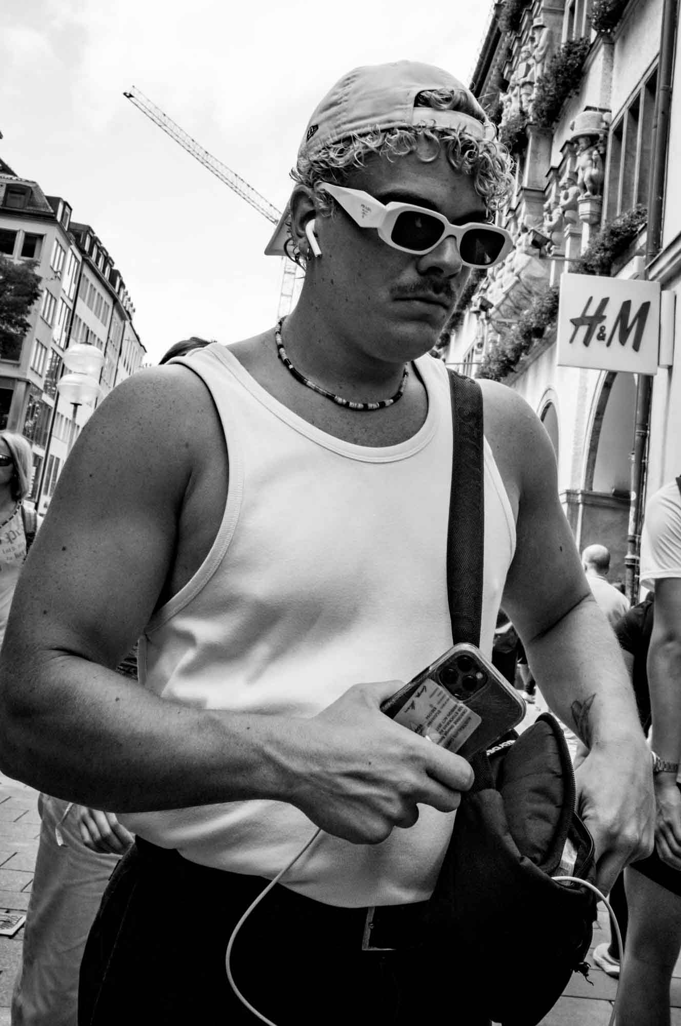 Man in a tank top and sunglasses walking in city, holding phone and bag, with H&M sign in background.