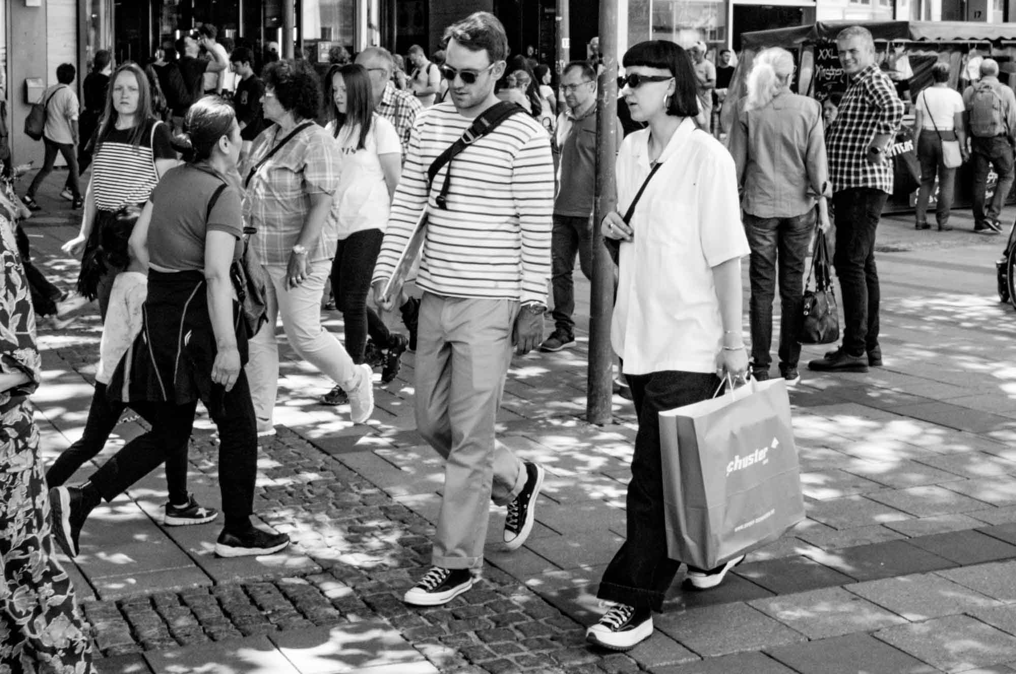 People walk down a busy city street on a sunny day, carrying shopping bags.