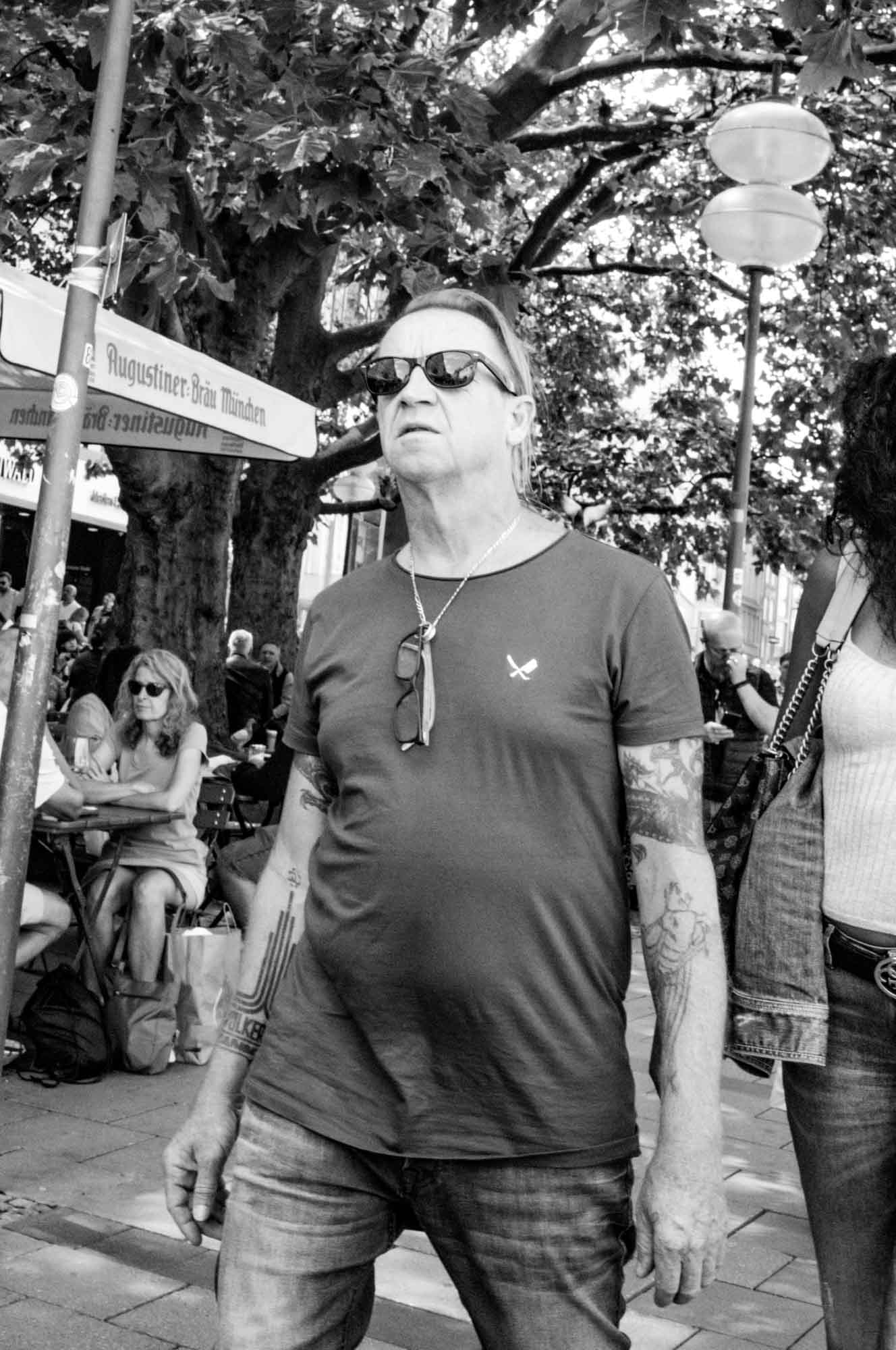 Man with tattoos wearing sunglasses, walking outdoors near cafe tables, surrounded by trees and people.