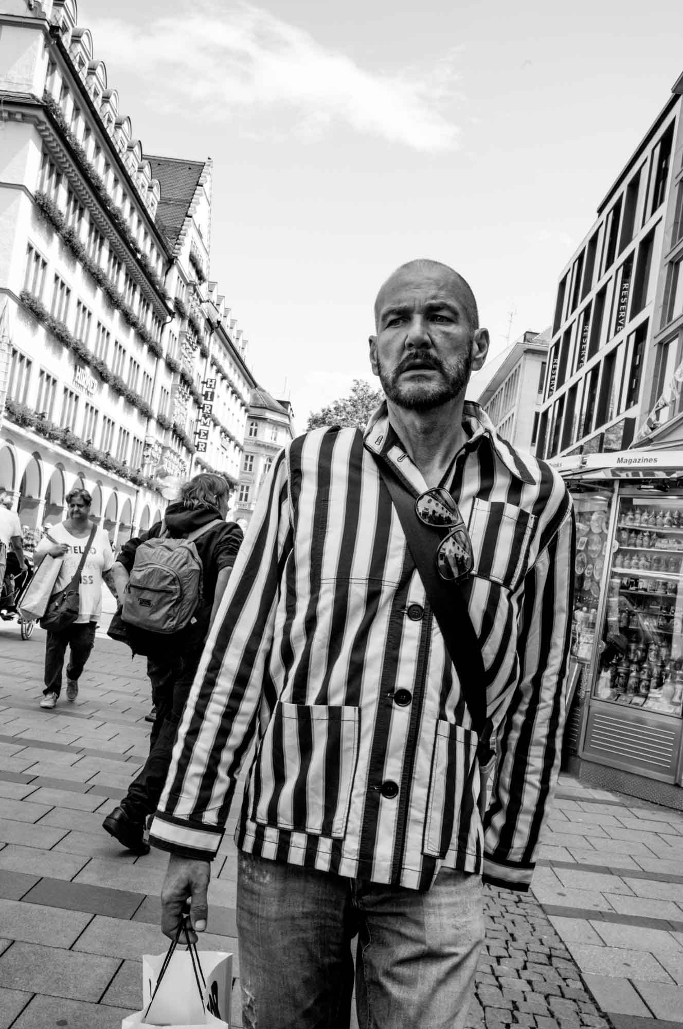 Man in striped jacket walking in city street, carrying shopping bags, black and white photo.
