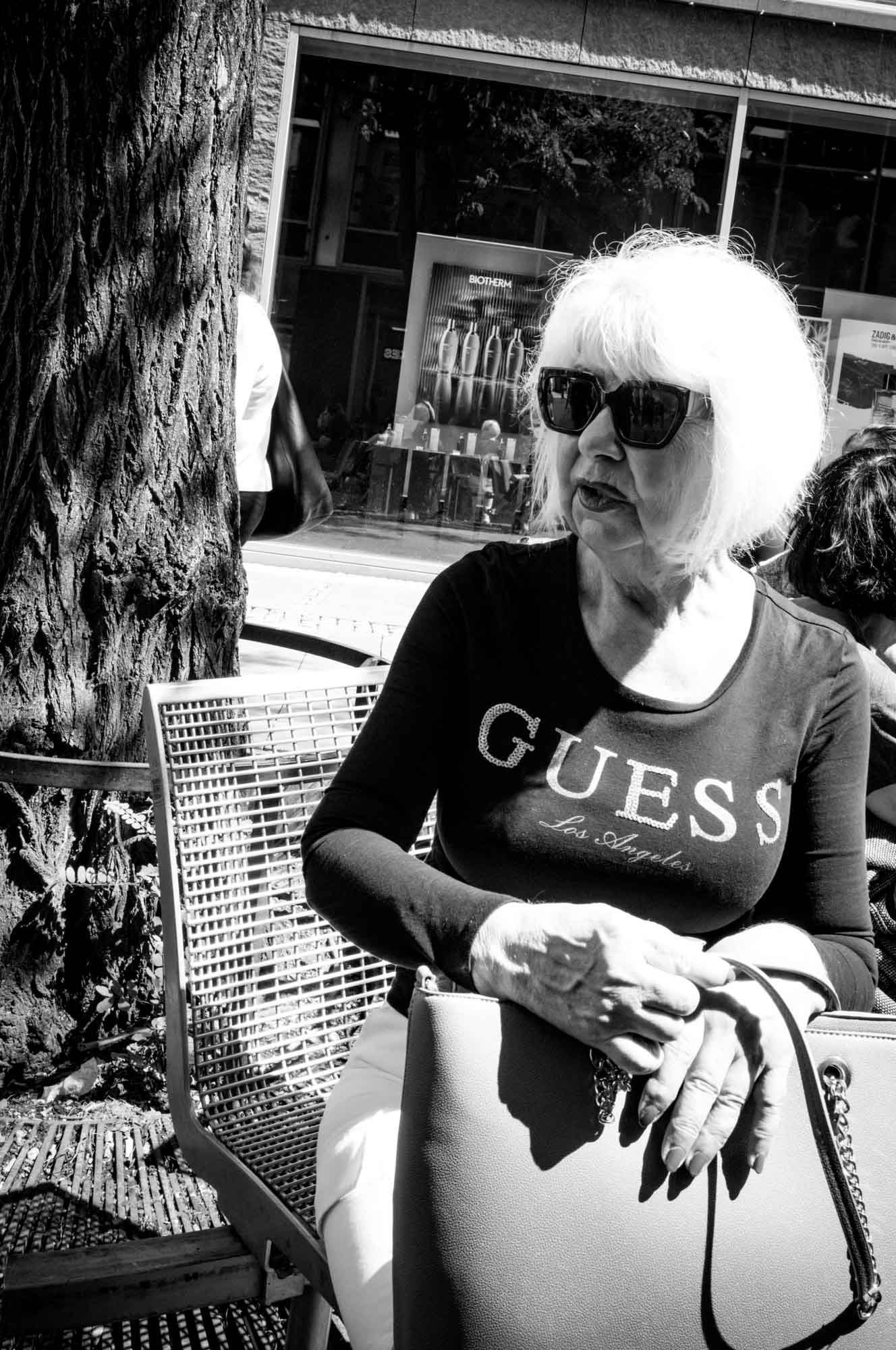 Woman in sunglasses and Guess shirt sits near a tree, holding a bag, in a sunny urban setting. Black and white photo.