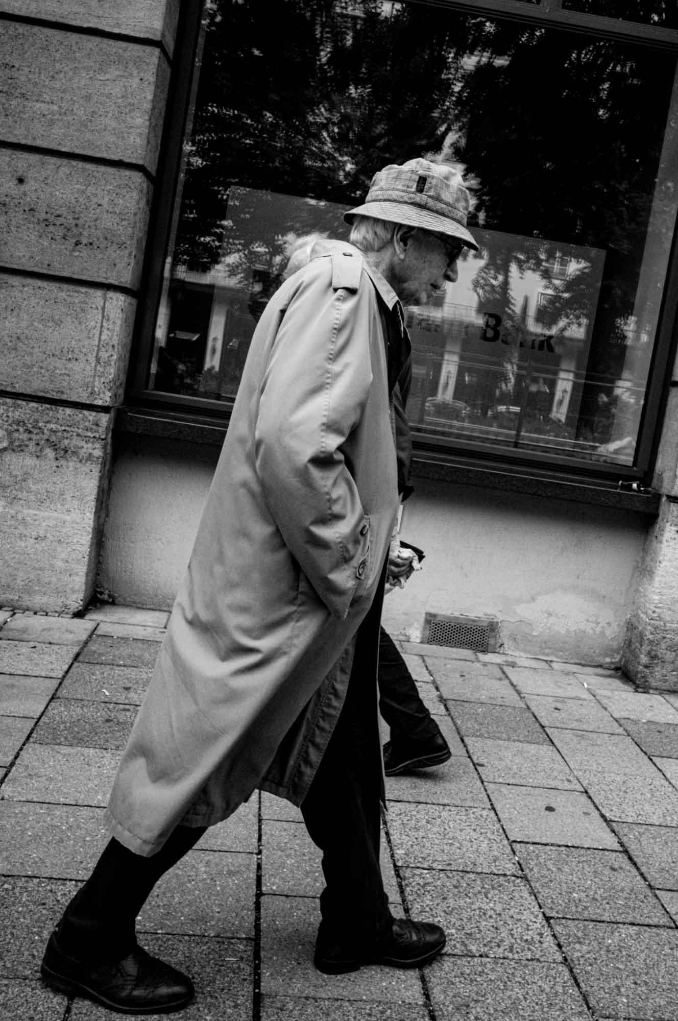 Older man in trench coat and hat walking on city sidewalk, black and white photo.