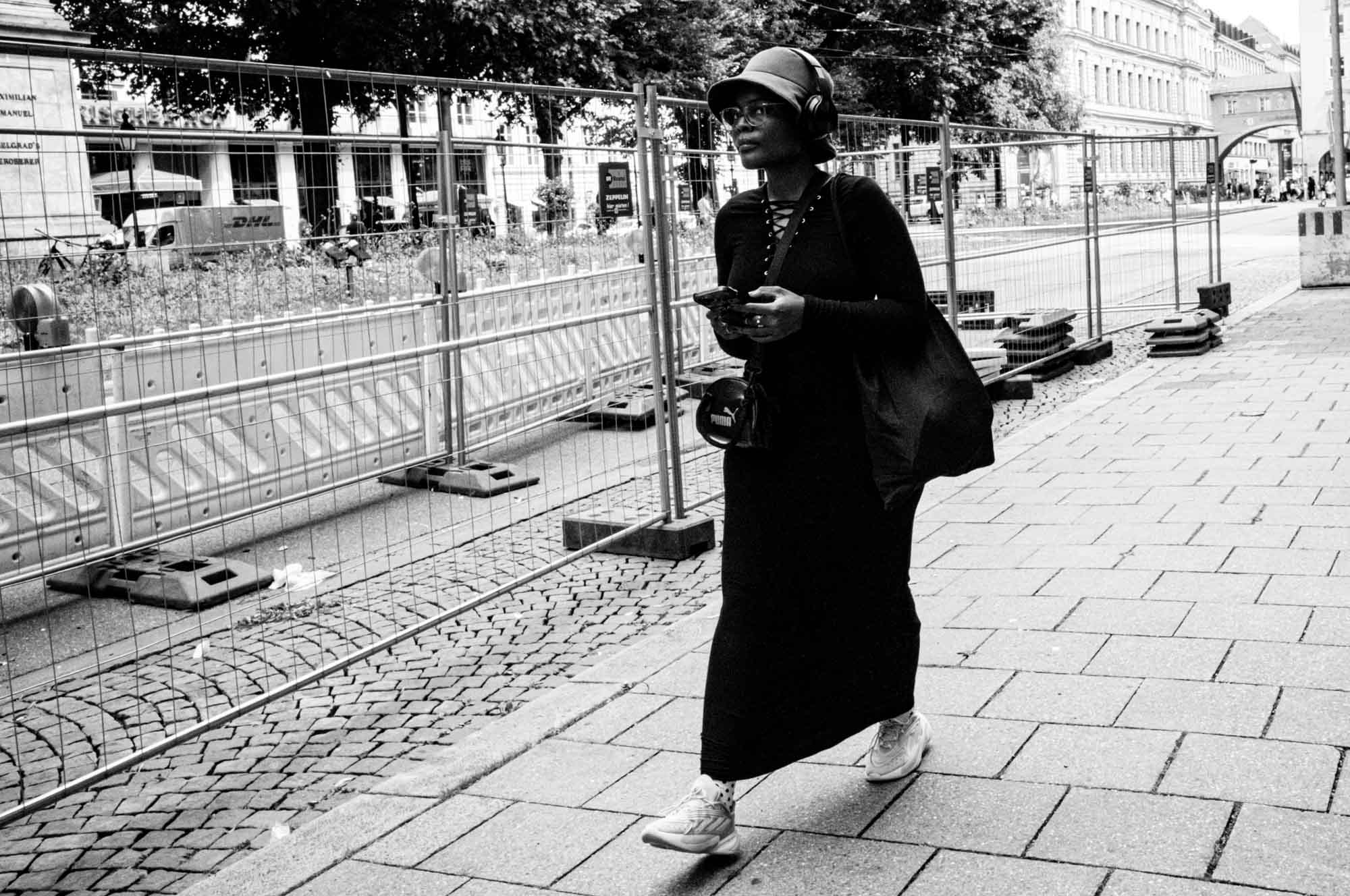 Person walking on city street with headphones, holding phone, near construction site. Black and white image.